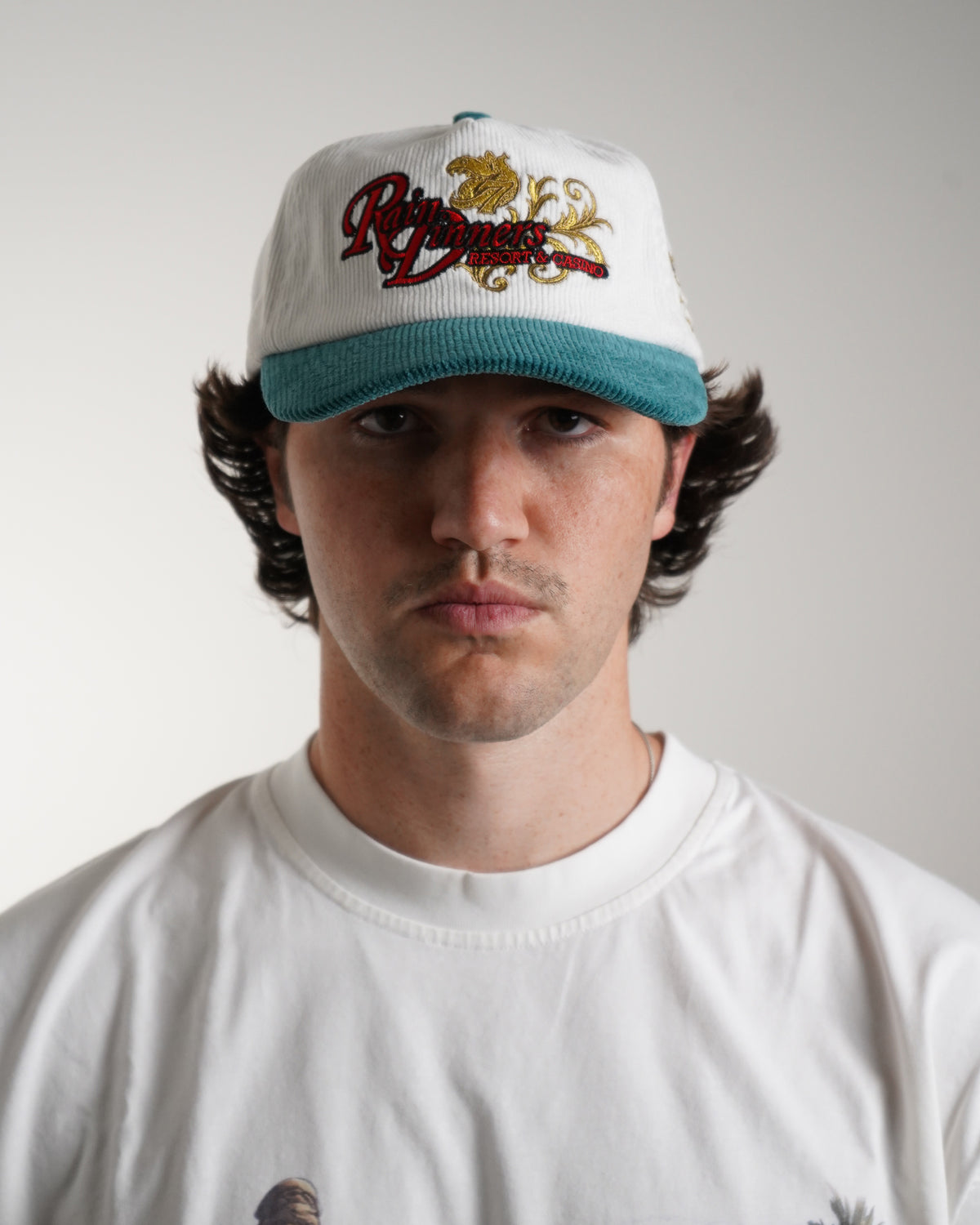 Model wearing the Rain Dinners Hat, a white corduroy cap with teal brim and embroidered red script, gold ornamental design and resort casino text, front view