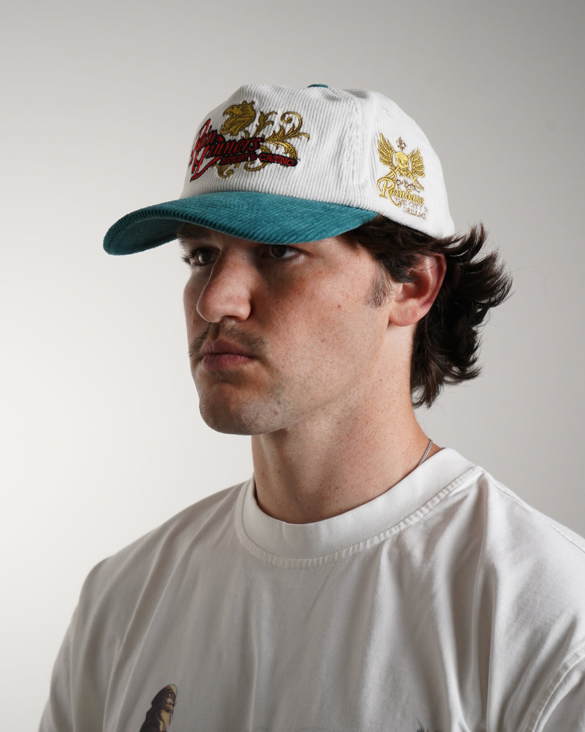 Model wearing the Rain Dinners Hat, a white corduroy cap with teal brim and gold and red embroidered graphics, angled side view