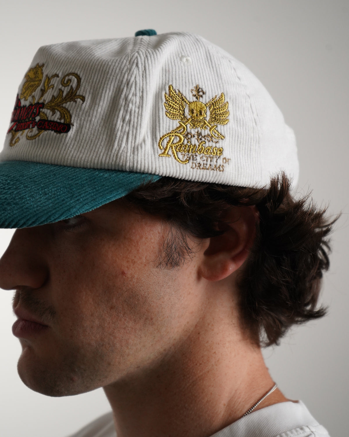 Side close-up on model of the Rain Dinners Hat, a white corduroy cap with teal brim, gold embroidered winged skull graphic and other embroidery