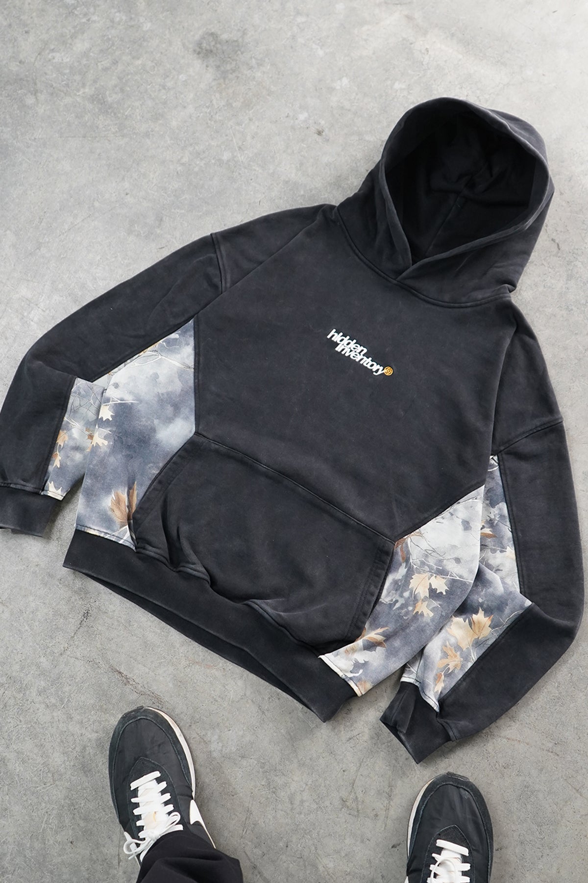Flat lay front view of the Hidden Inventory Hoodie in washed black with gray leaf camouflage side panels and small embroidered chest text