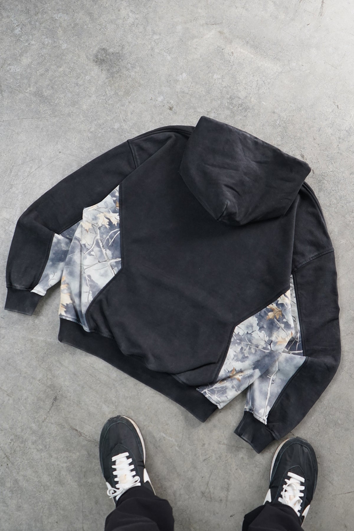Back flat lay of the faded black Hidden Inventory Hoodie with hood, gray-white leafy camouflage side panels, and a person's black sneakers below