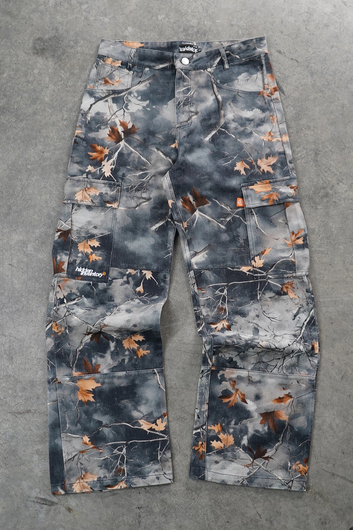 A pair of cargo pants featuring a dark gray and black cloudy camouflage pattern with scattered brown and orange autumn leaves and branches, laid flat on a gray concrete surface.