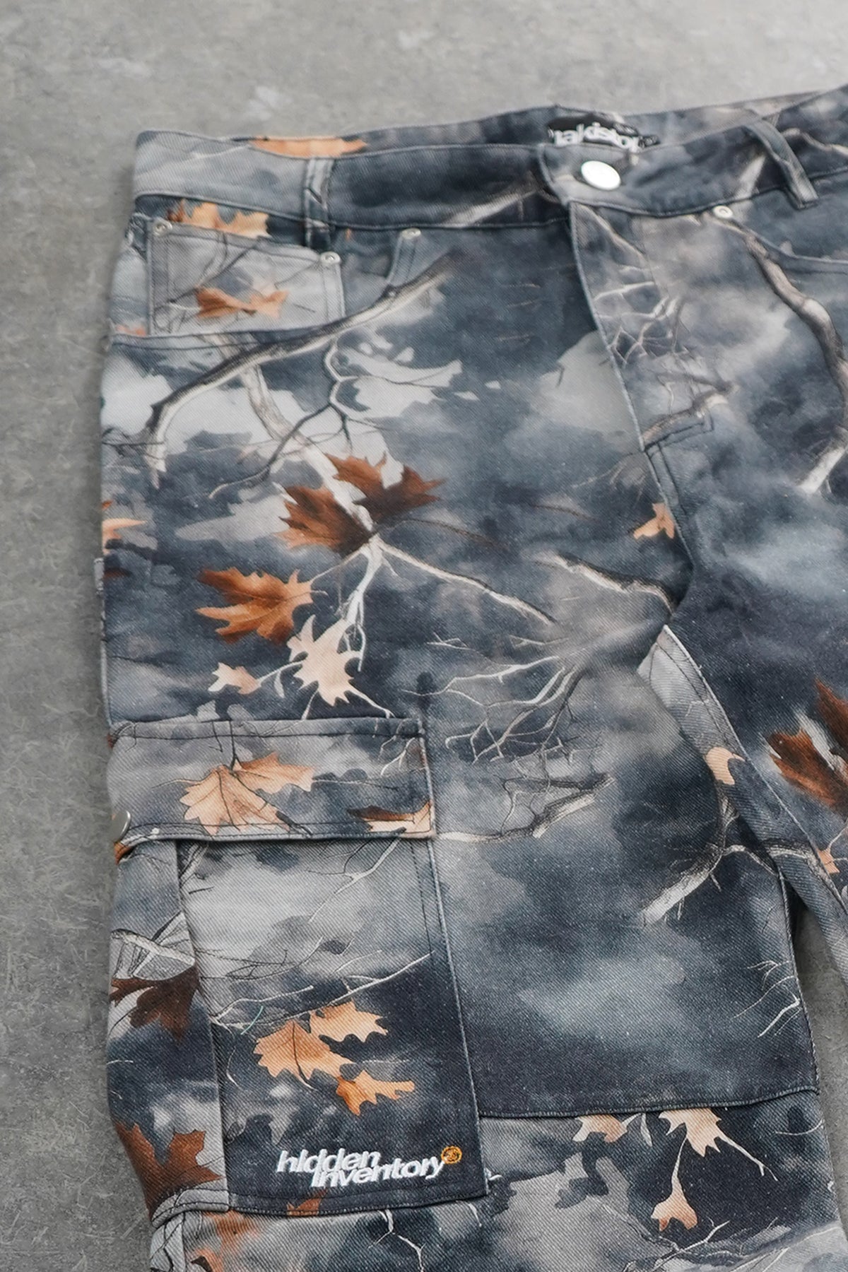 Close-up of a pair of cargo pants laid flat on a gray surface, featuring a dark gray and black bark camouflage pattern with brown autumn leaves and a visible 'hikkit' brand patch near the lower pocket.