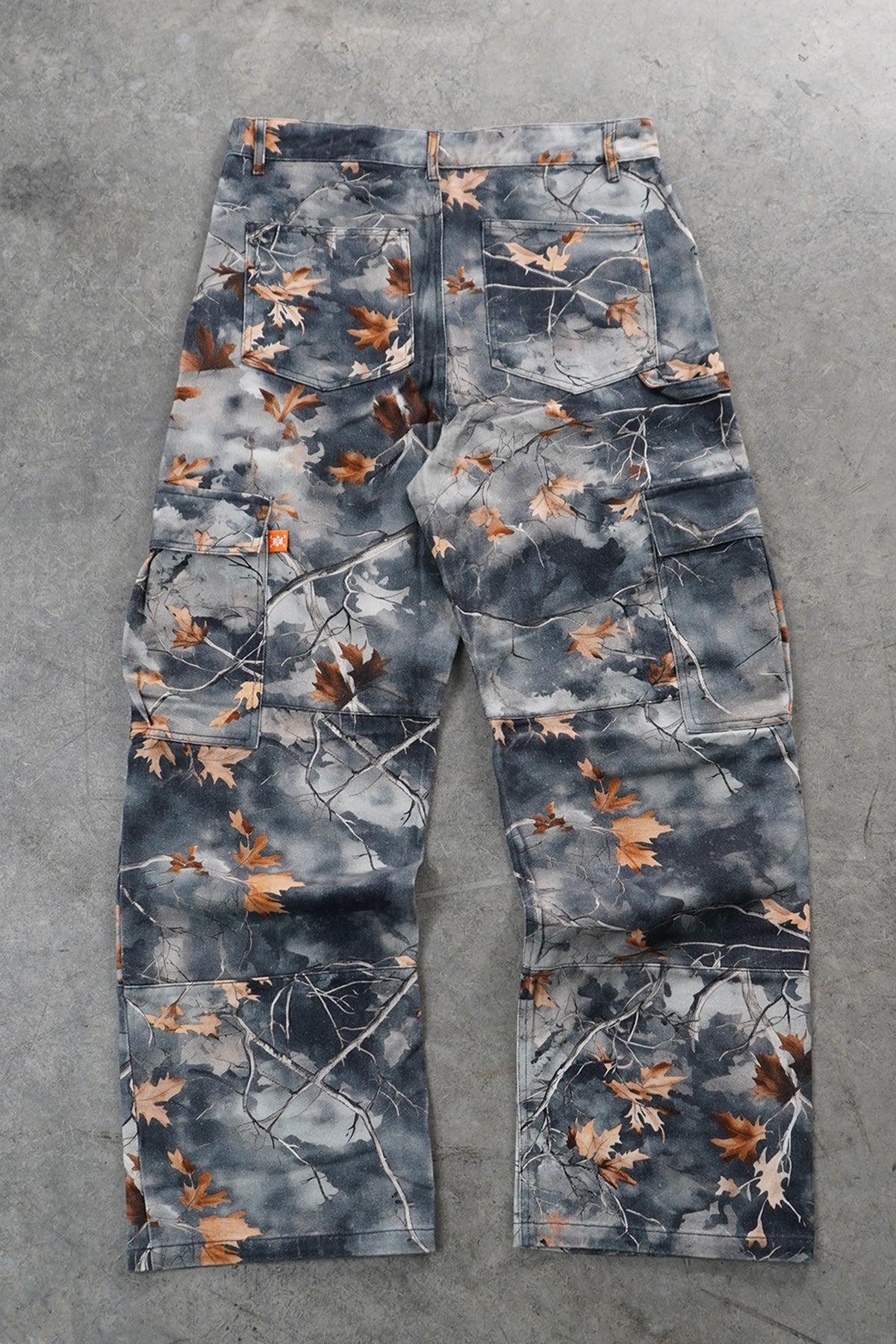 A pair of cargo pants featuring a gray and blue-gray tree branch camouflage pattern with scattered brown leaves, laid flat on a gray concrete surface.