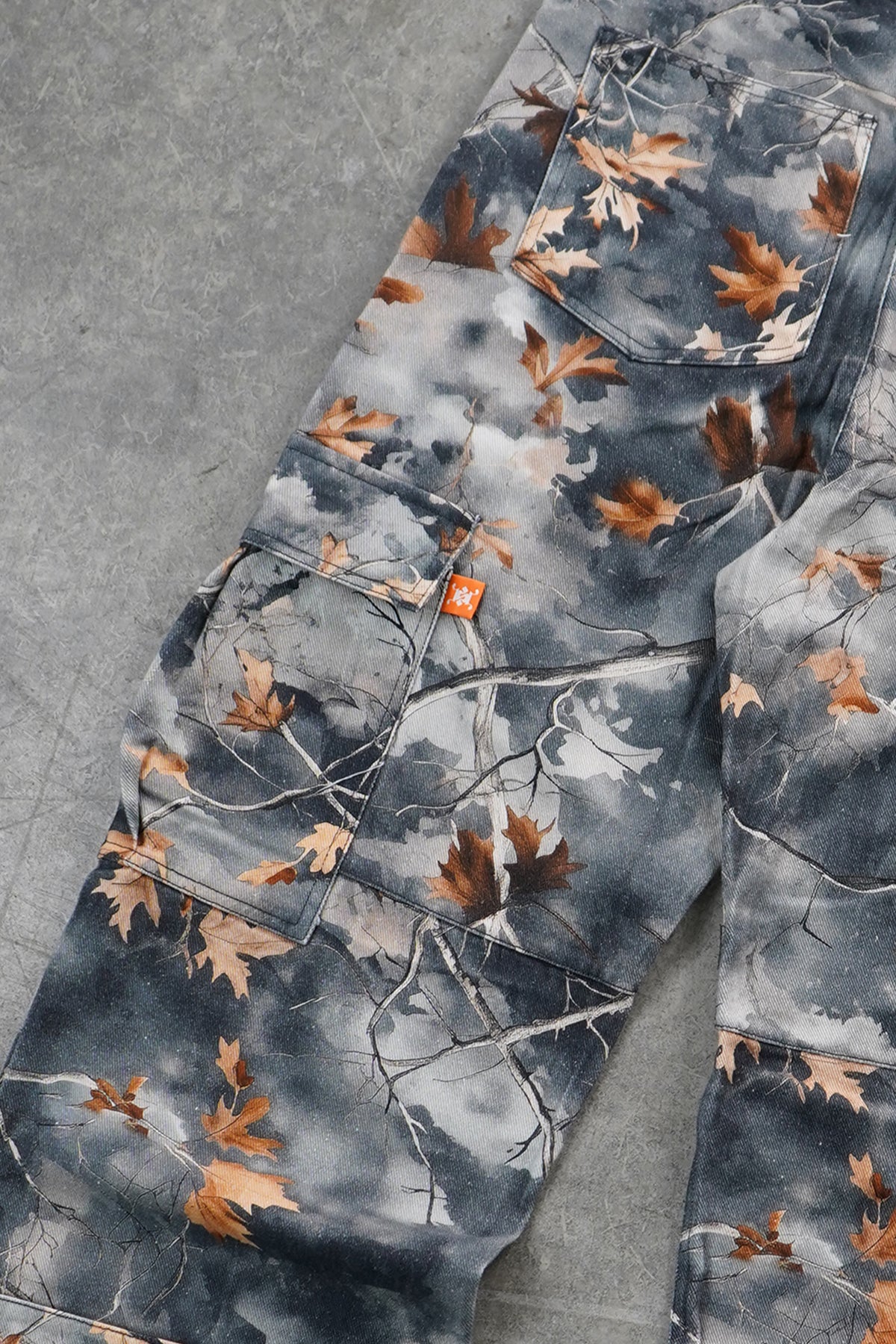 A close-up of camouflage pants featuring a gray, white, and brown leaf-and-branch woodland pattern, laid flat on a gray textured surface.