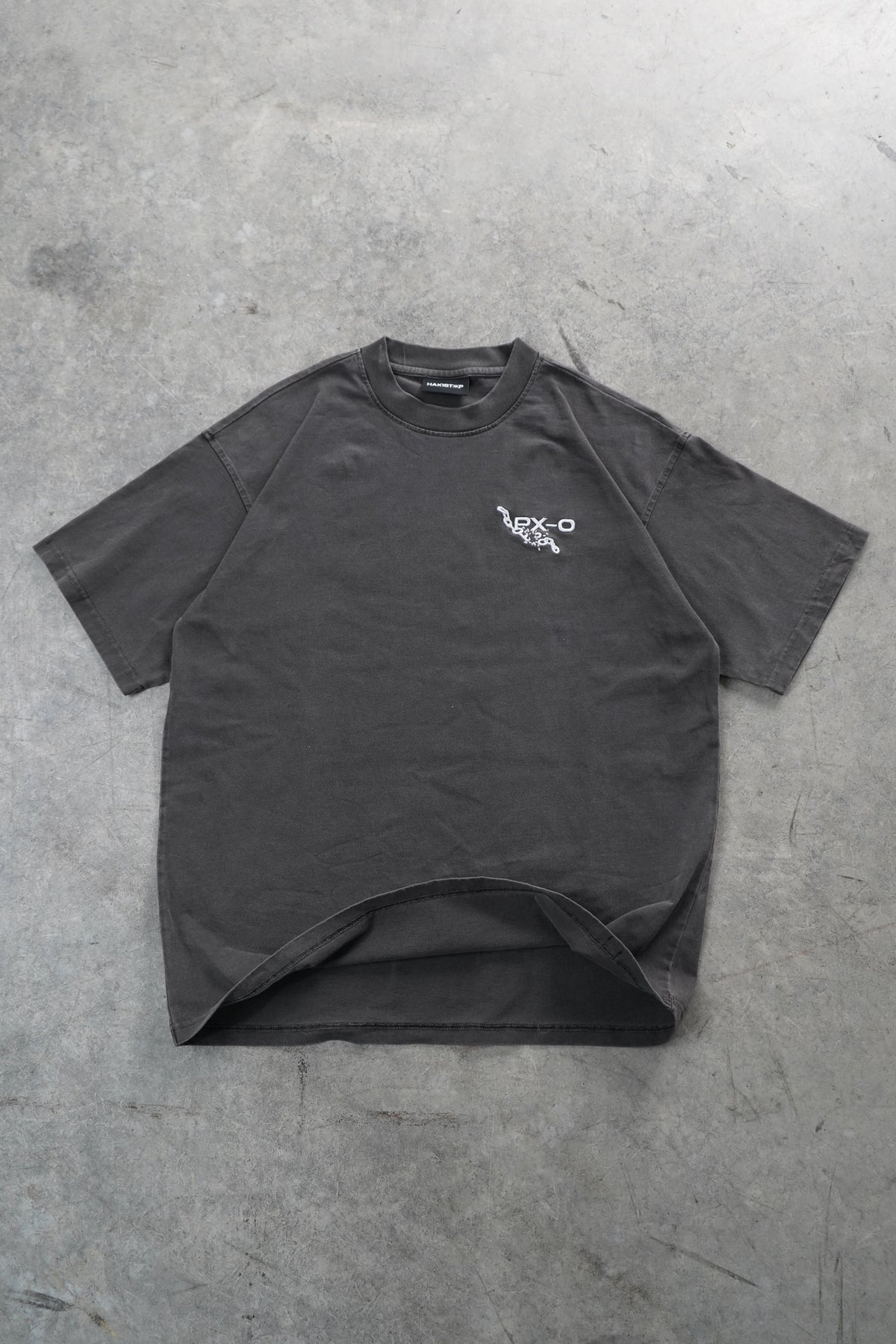 A dark gray oversized short-sleeve t-shirt with a small white graphic on the left chest, laid flat on a textured gray concrete floor.