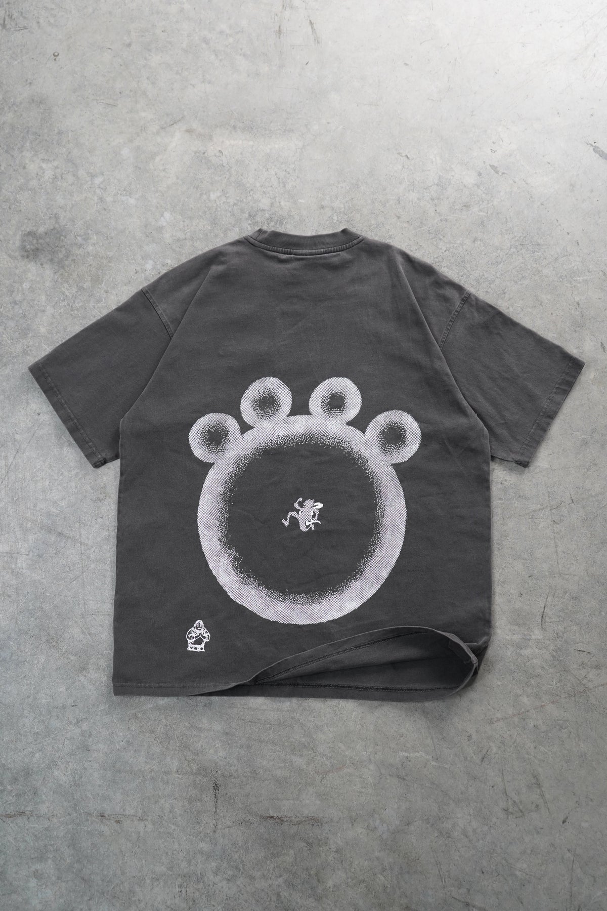 A dark gray washed t-shirt laid flat on a textured gray concrete surface, featuring a large white circular graphic with three circular details on top resembling a bear or paw print.
