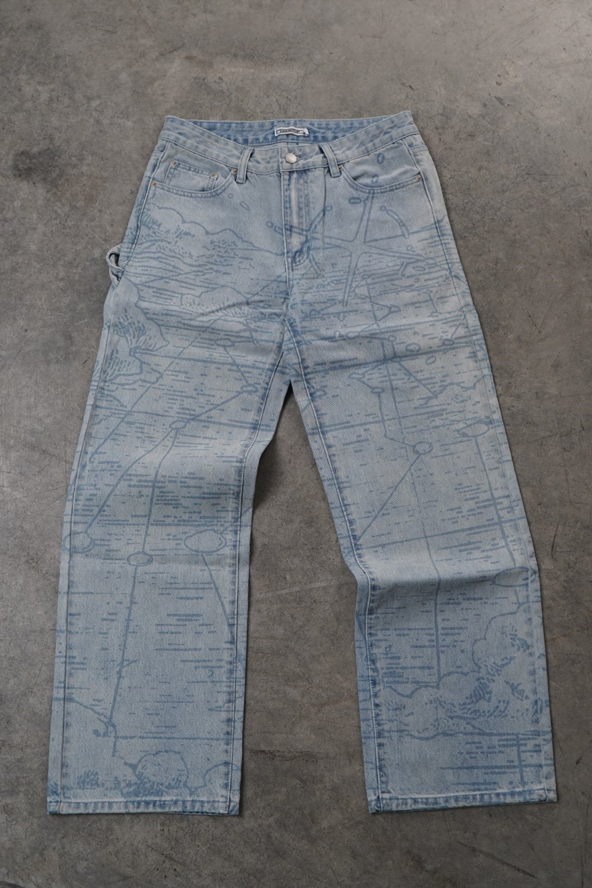 Light blue denim jeans with a faint blue map print, laid flat on a gray concrete surface.