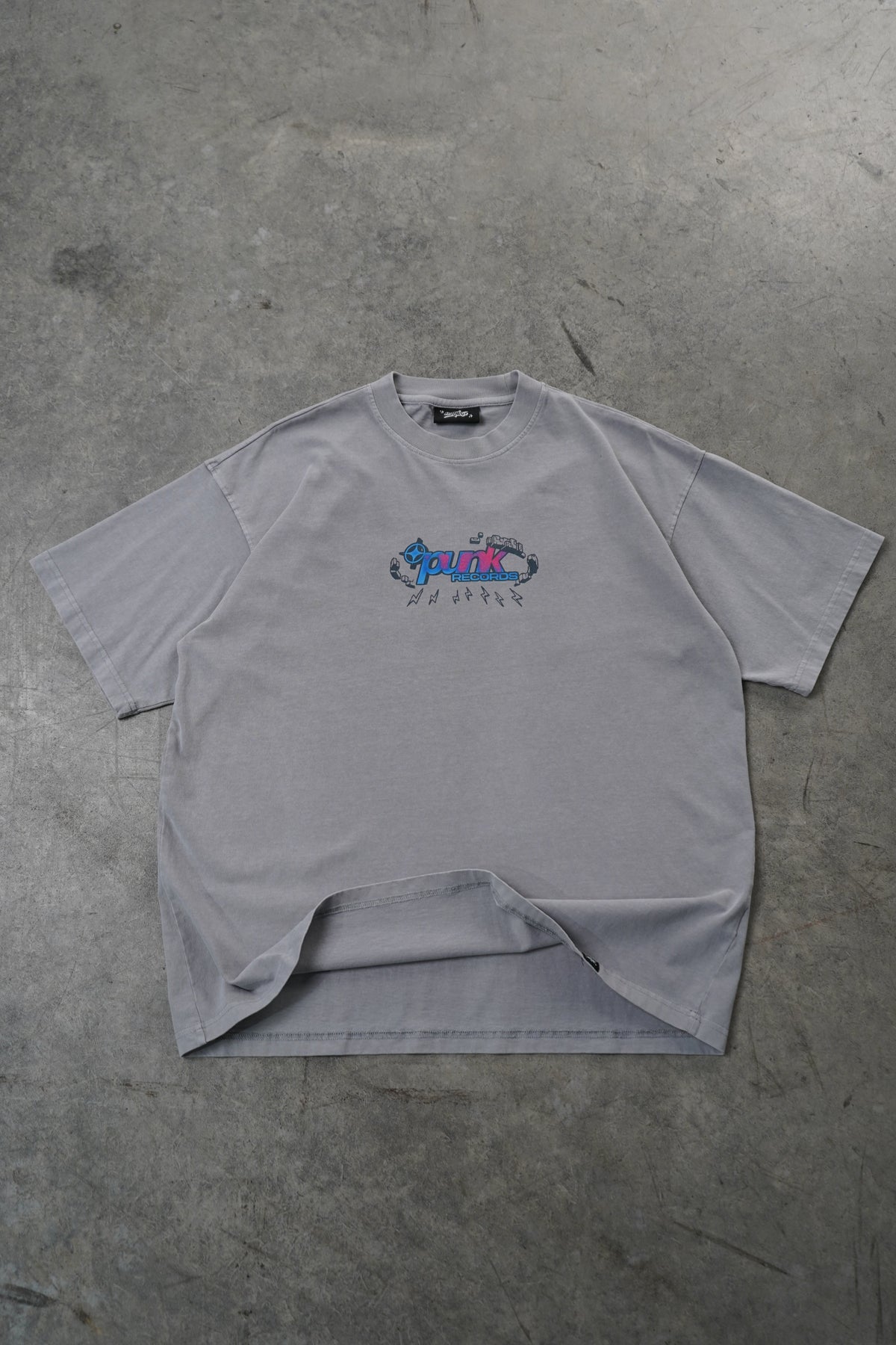 A light gray oversized short-sleeve t-shirt with a small colorful graphic on the left chest, laid flat on a textured gray concrete floor.