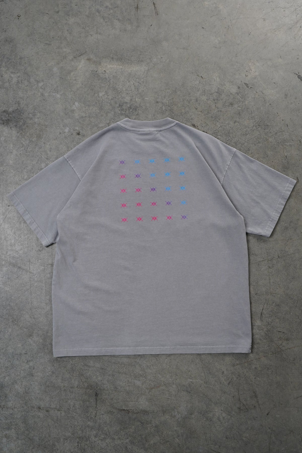 A light gray oversized long-sleeve t-shirt laid flat on a textured gray concrete floor. The shirt features a small grid of colorful symbols printed on the chest, with pink, purple, and blue star-like marks.