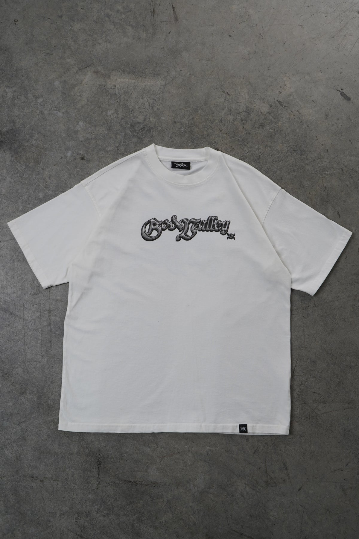 A white short-sleeve t-shirt with a black vertical graphic print on the right chest, laid flat on a gray concrete floor. The brand name 'God Valley' is visible in the graphic.