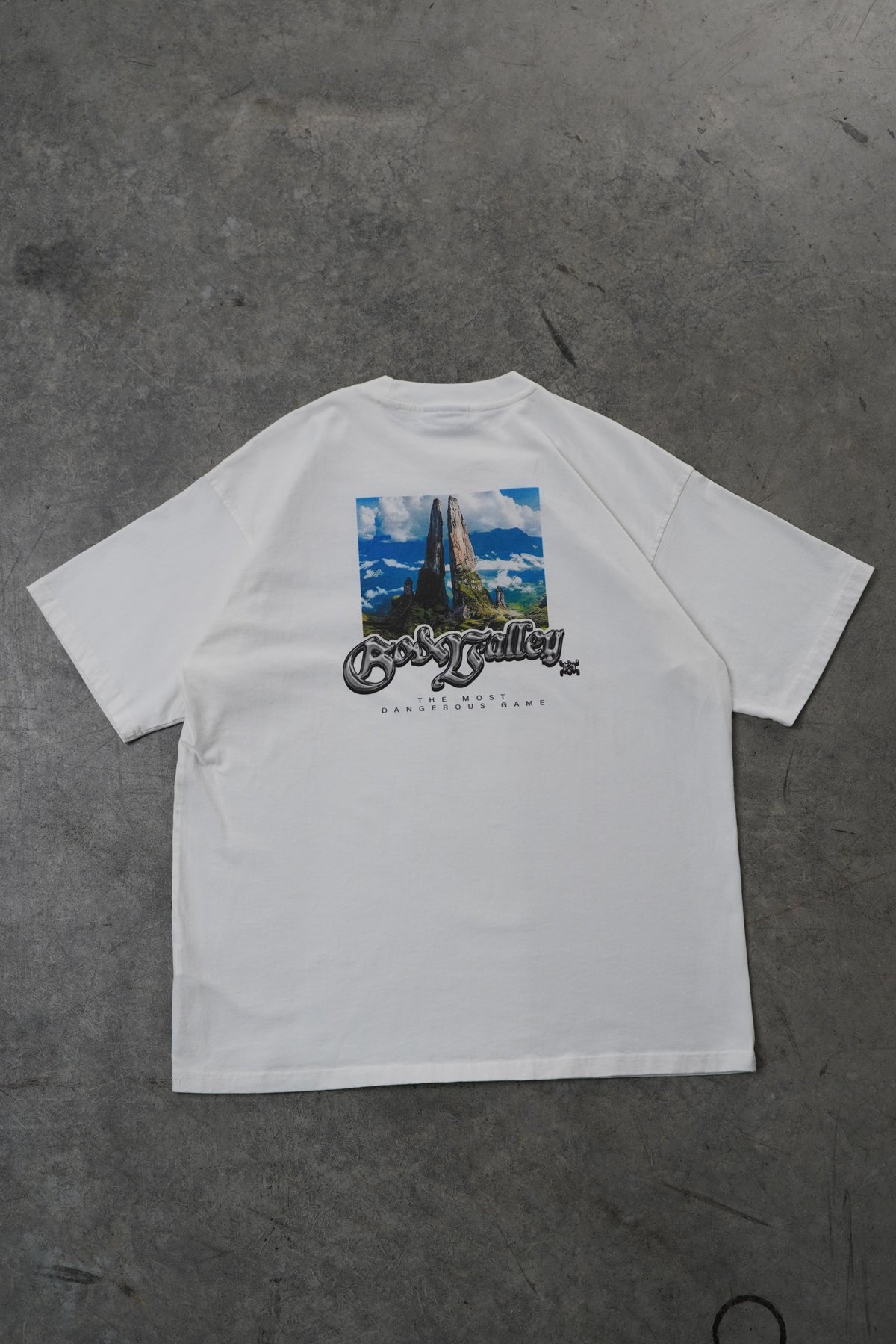 A white short-sleeve t-shirt laid flat on a textured gray concrete floor, featuring a graphic print on the front with a blue sky and mountain scene alongside stylized text.