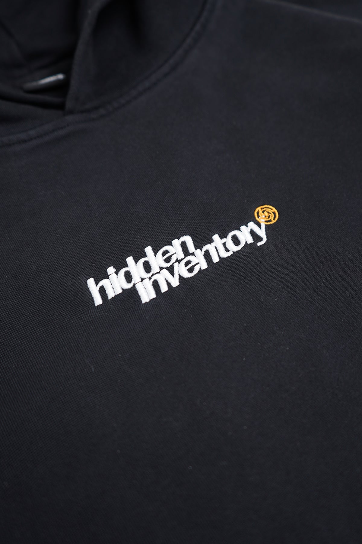 Close-up of the black Hidden Inventory Hoodie chest, with white embroidered text and a small orange spiral emblem beside it