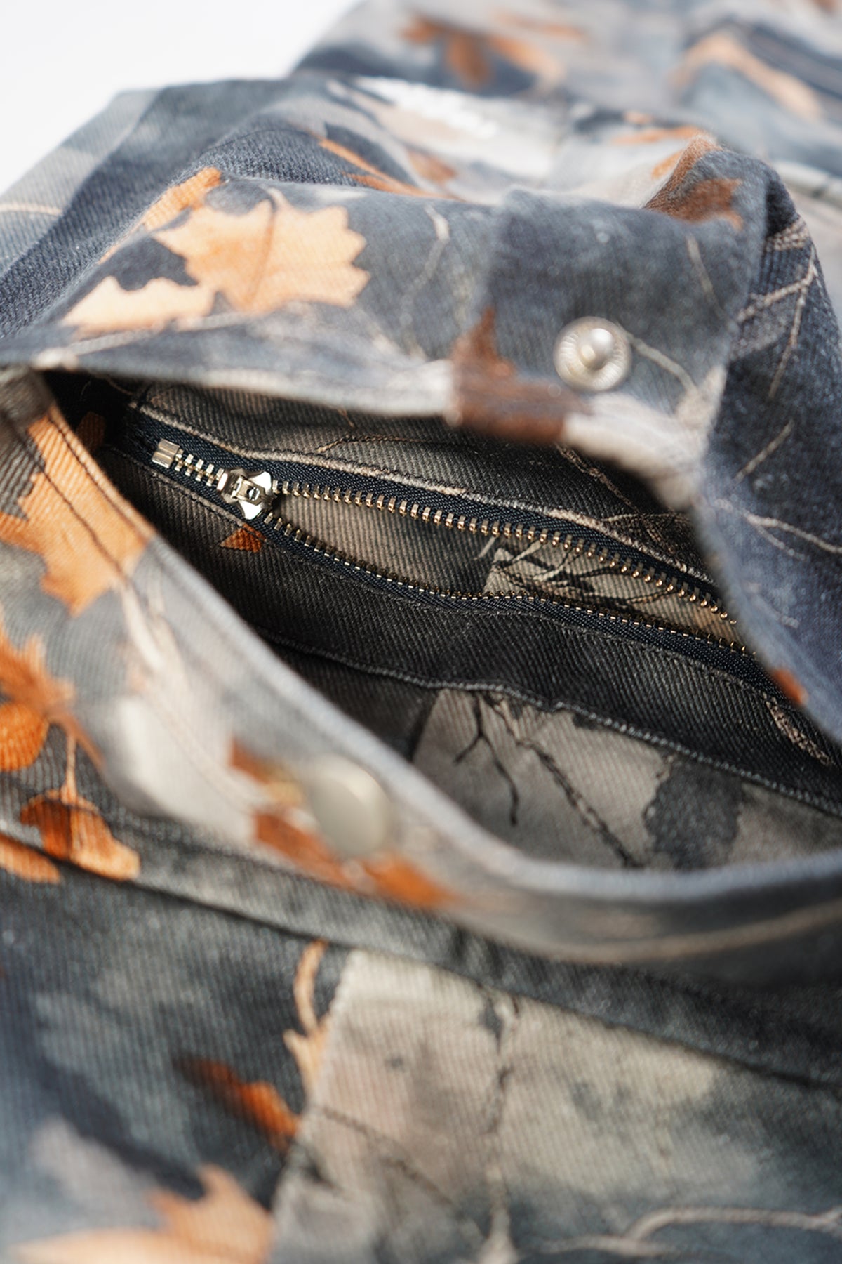 Close-up of a camouflage-patterned fabric item, likely a bag or pouch, showing a dark zipper opening with a leaf and branch print in muted blue, gray, and brown tones.