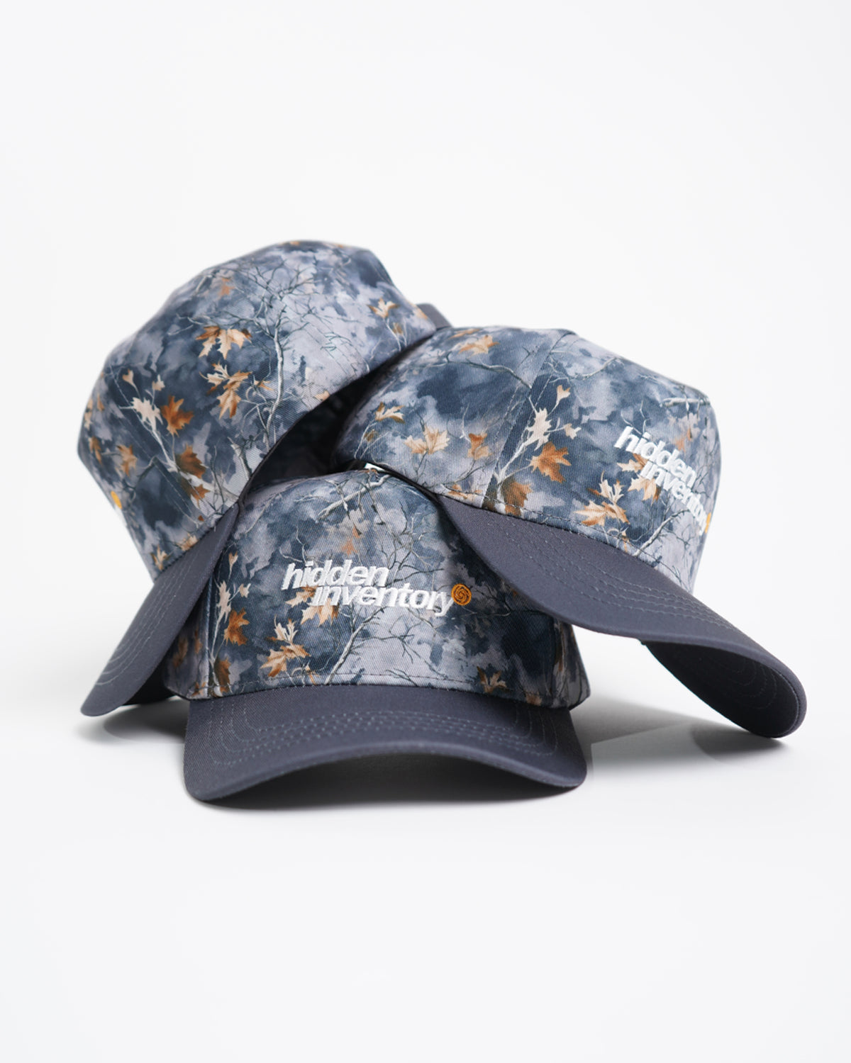 Stack of three Hidden Inventory Hat caps in blue-gray autumn leaf camo with dark gray brims and white embroidered text, front and side views
