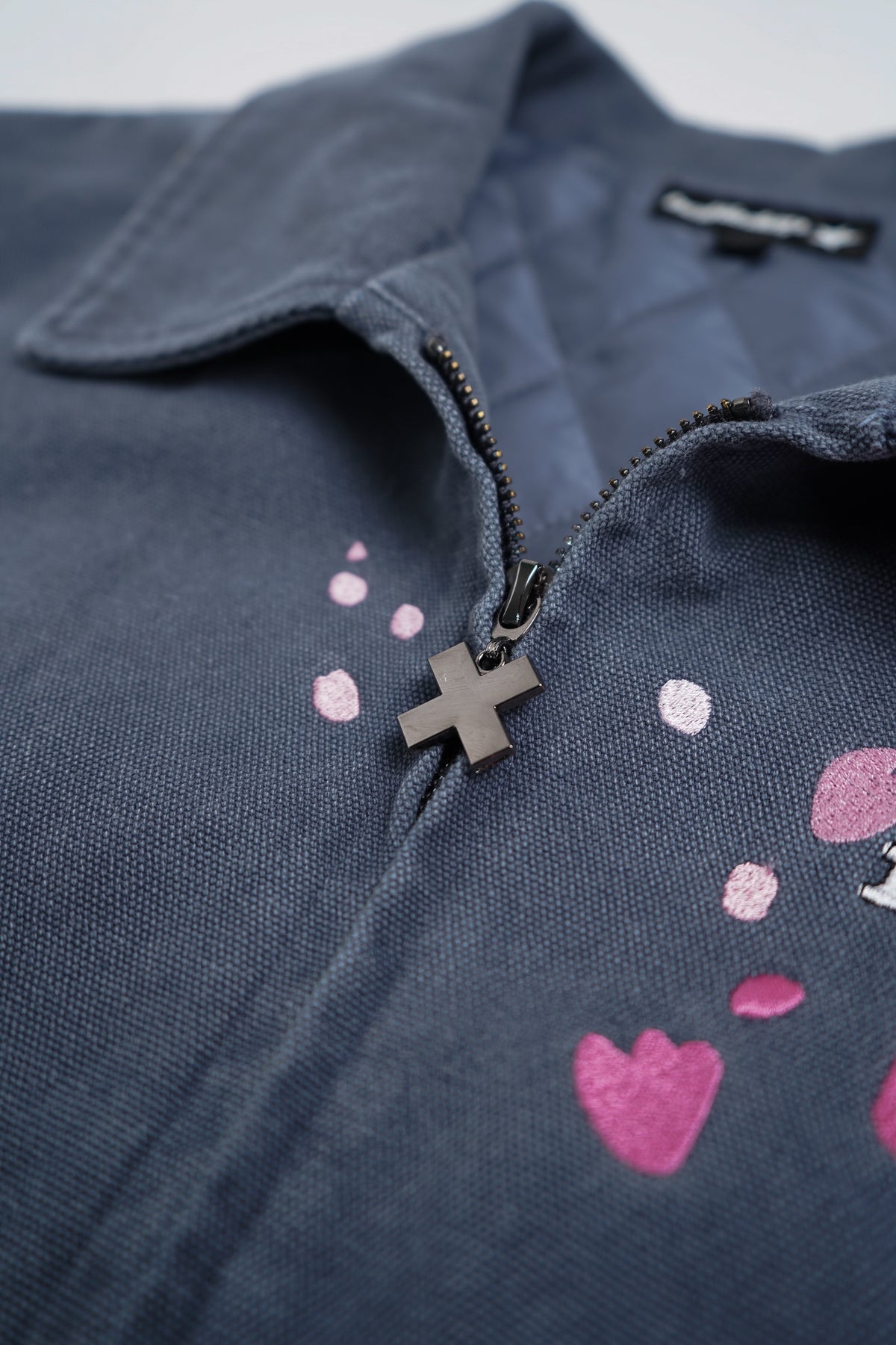 Close-up of a dark navy blue zip-up jacket or overshirt with a black zipper and a small metallic cross charm. The fabric features pink glittery heart and dot patterns.