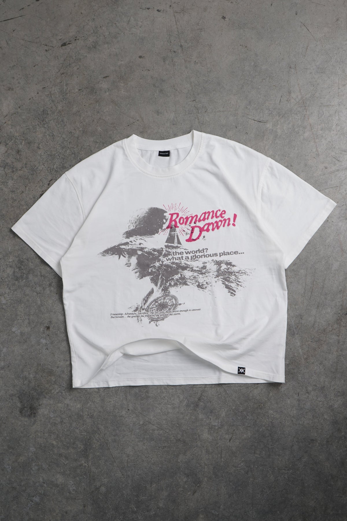 A white oversized graphic T-shirt laid flat on a textured gray concrete floor. The shirt features a black and gray tree illustration with red script text on the front.