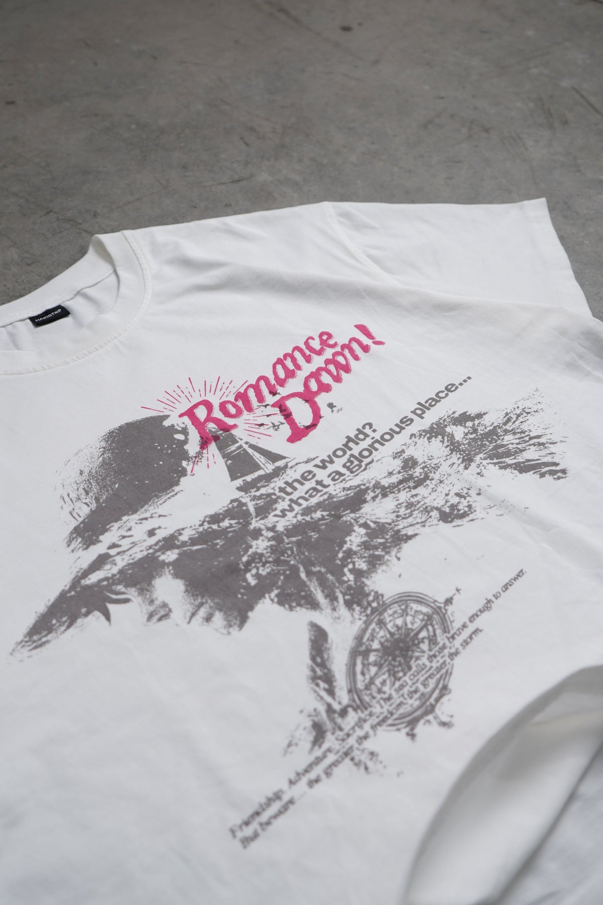 A white graphic t-shirt laid flat on a gray concrete floor. The shirt features a black and pink graphic print with the text 'Romance Dawn!' and a scenic illustration.