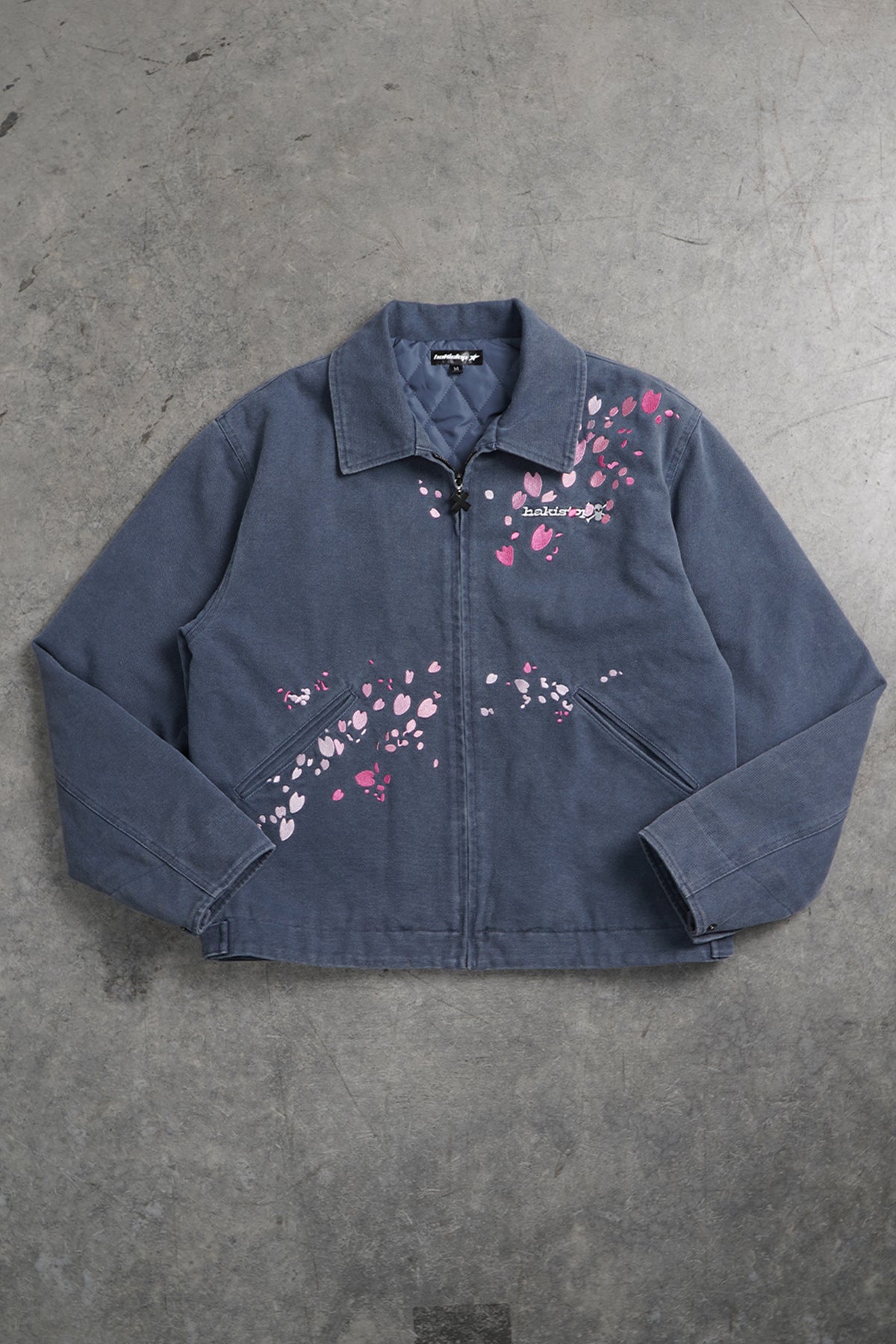A dark blue-gray jacket with pink cherry blossom prints, laid flat on a textured gray concrete floor.