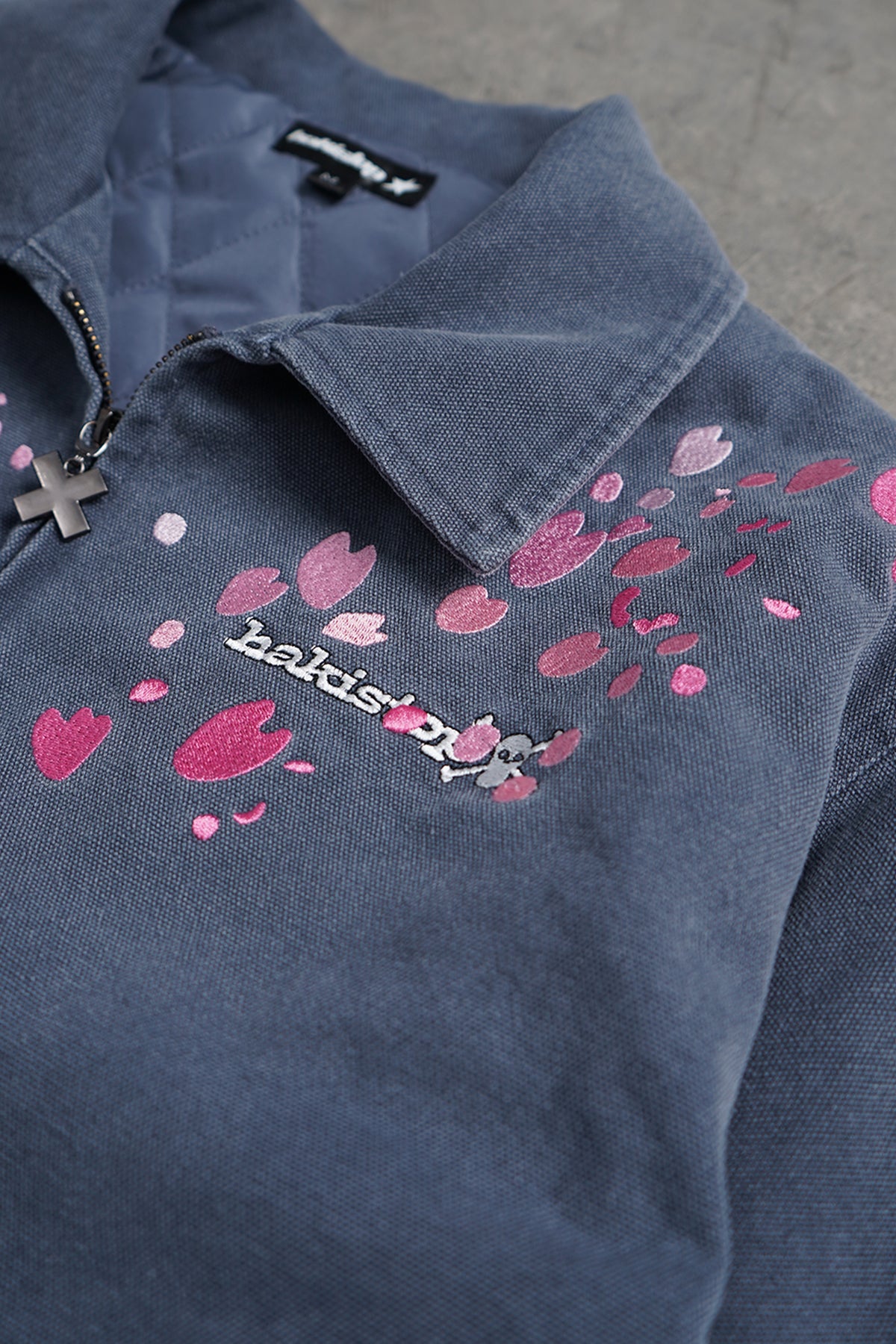 Close-up of a dark blue-gray collared pullover with a partial zipper, pink heart and flower embroidery, and a small skull-and-crossbones detail on the chest.