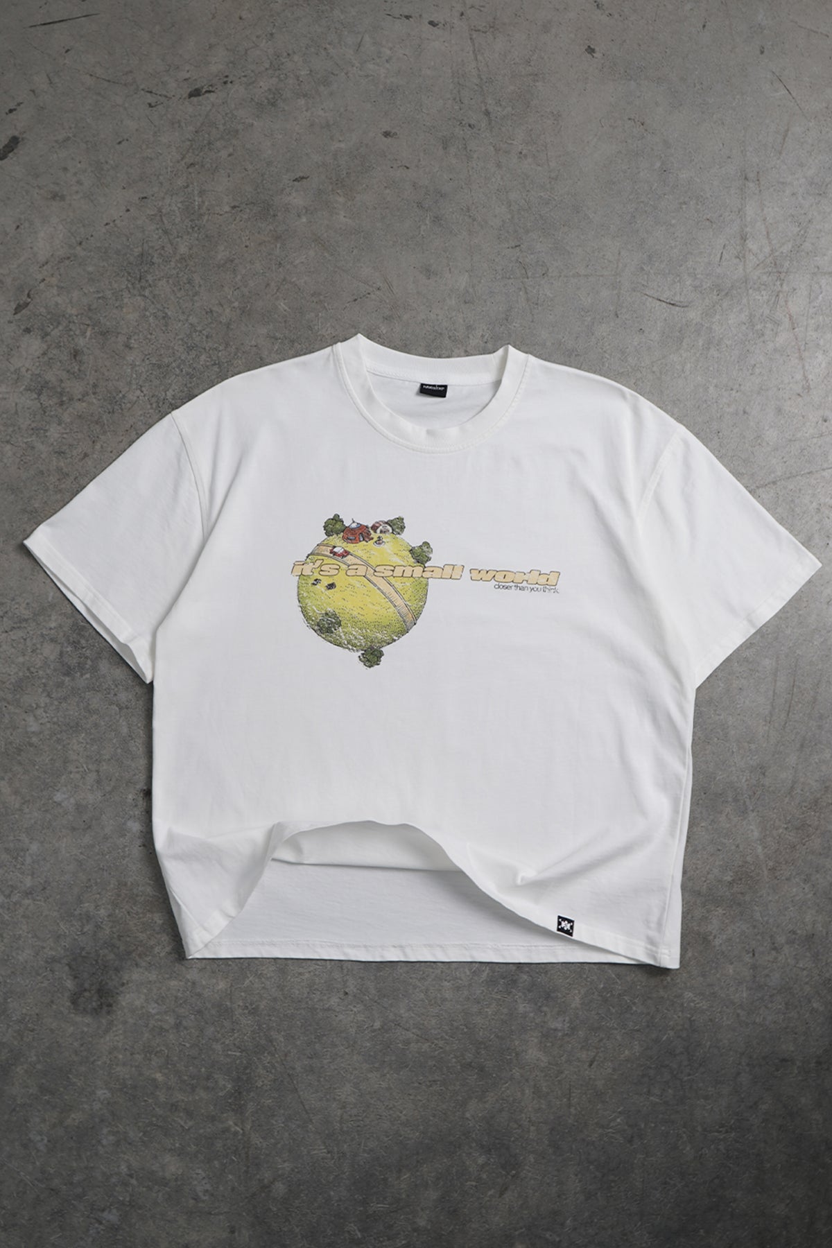 Flat lay front view of the white Small World Tee with a small green planet graphic with tiny houses, trees and a road, plus printed text