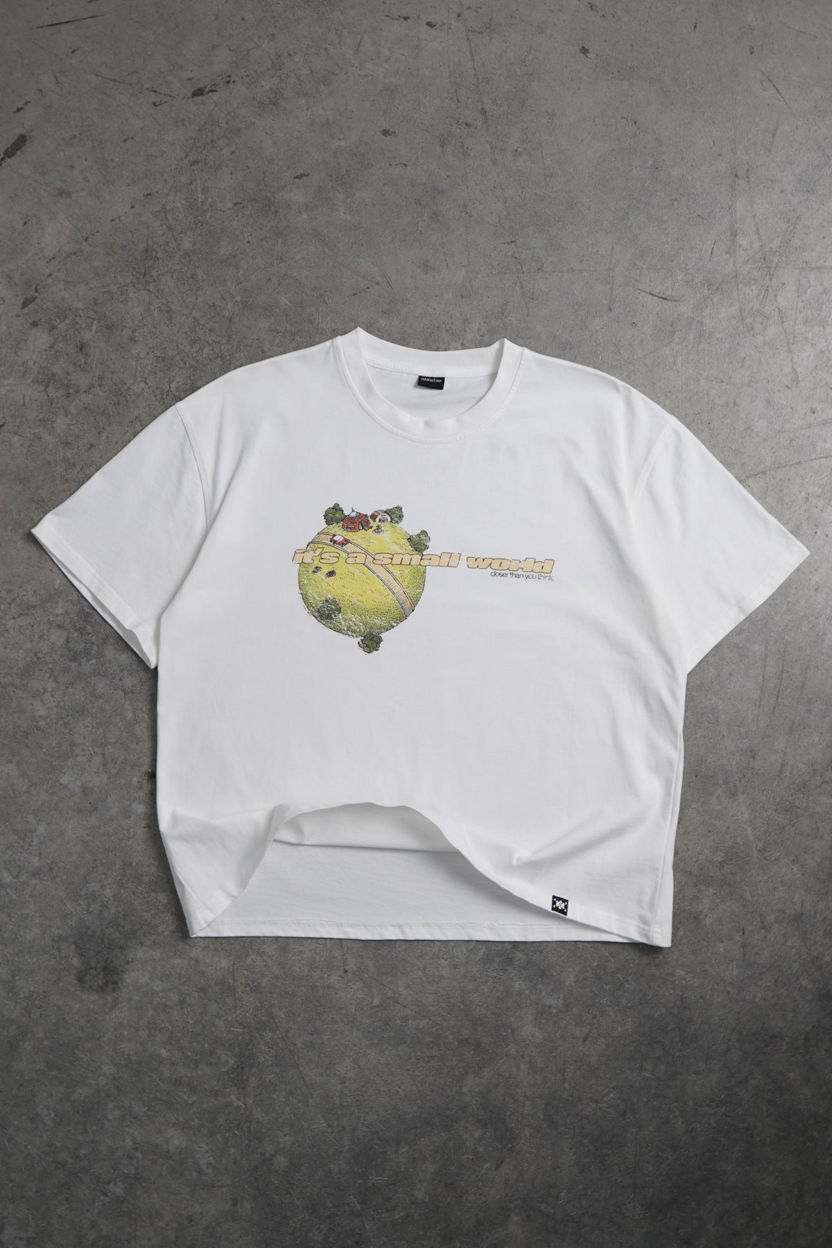 Flat lay front view of the white Small World Tee from the Dragonball 40th Anniversary Tee Bundle, with a small green planet graphic and printed text