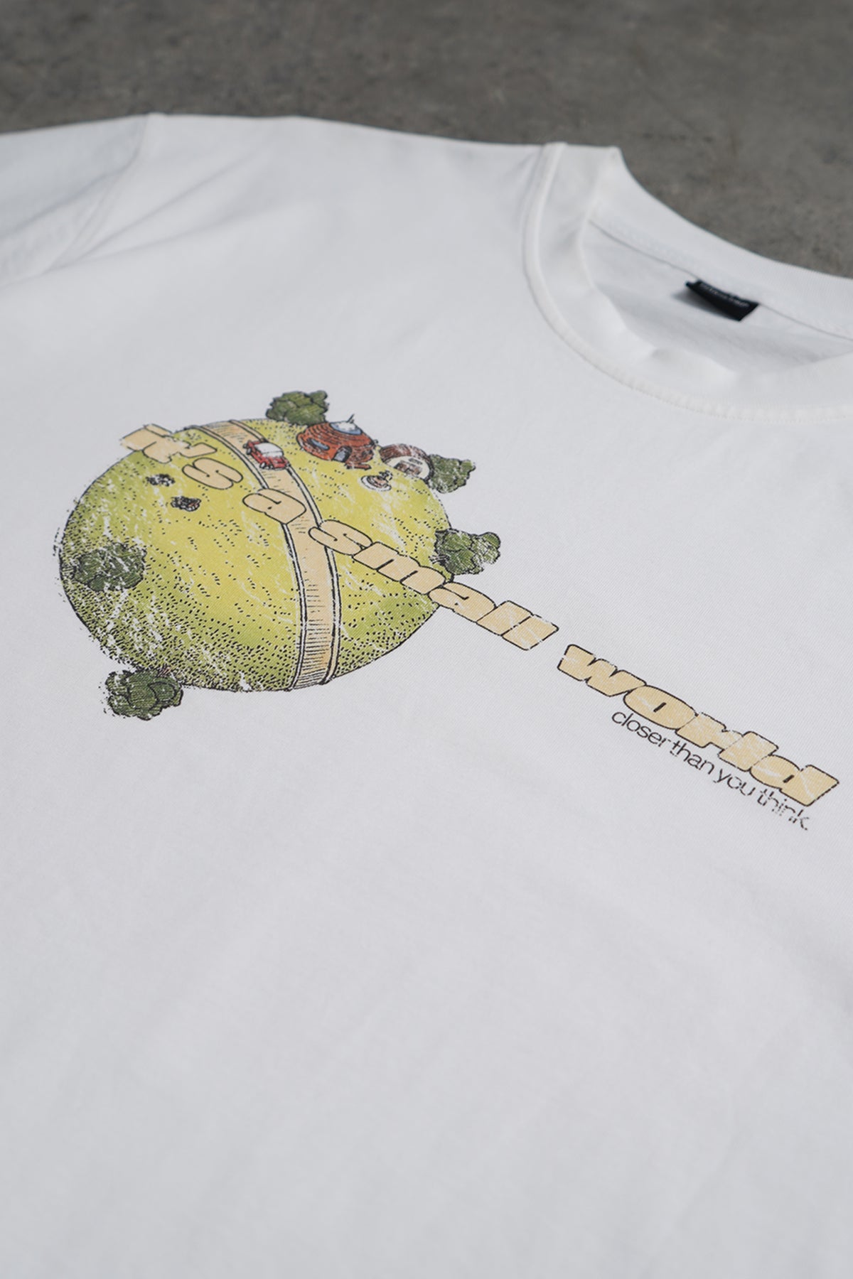 Angled close-up of the white Small World Tee on a concrete surface, with a small green planet graphic, cream lettering reading "It's a small world" and smaller text below