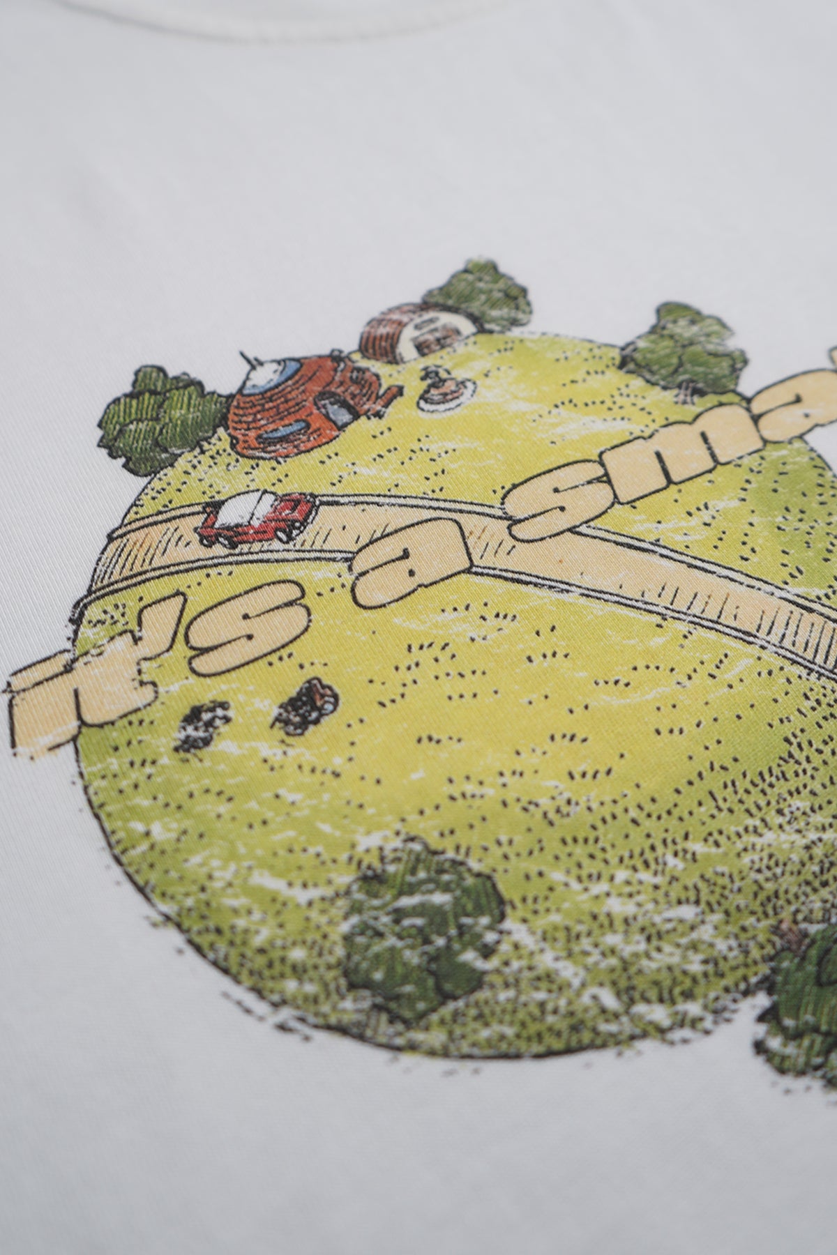 Close-up of the white Small World Tee front graphic: a small round green planet with a tan road, red car, round houses, trees and cropped printed text