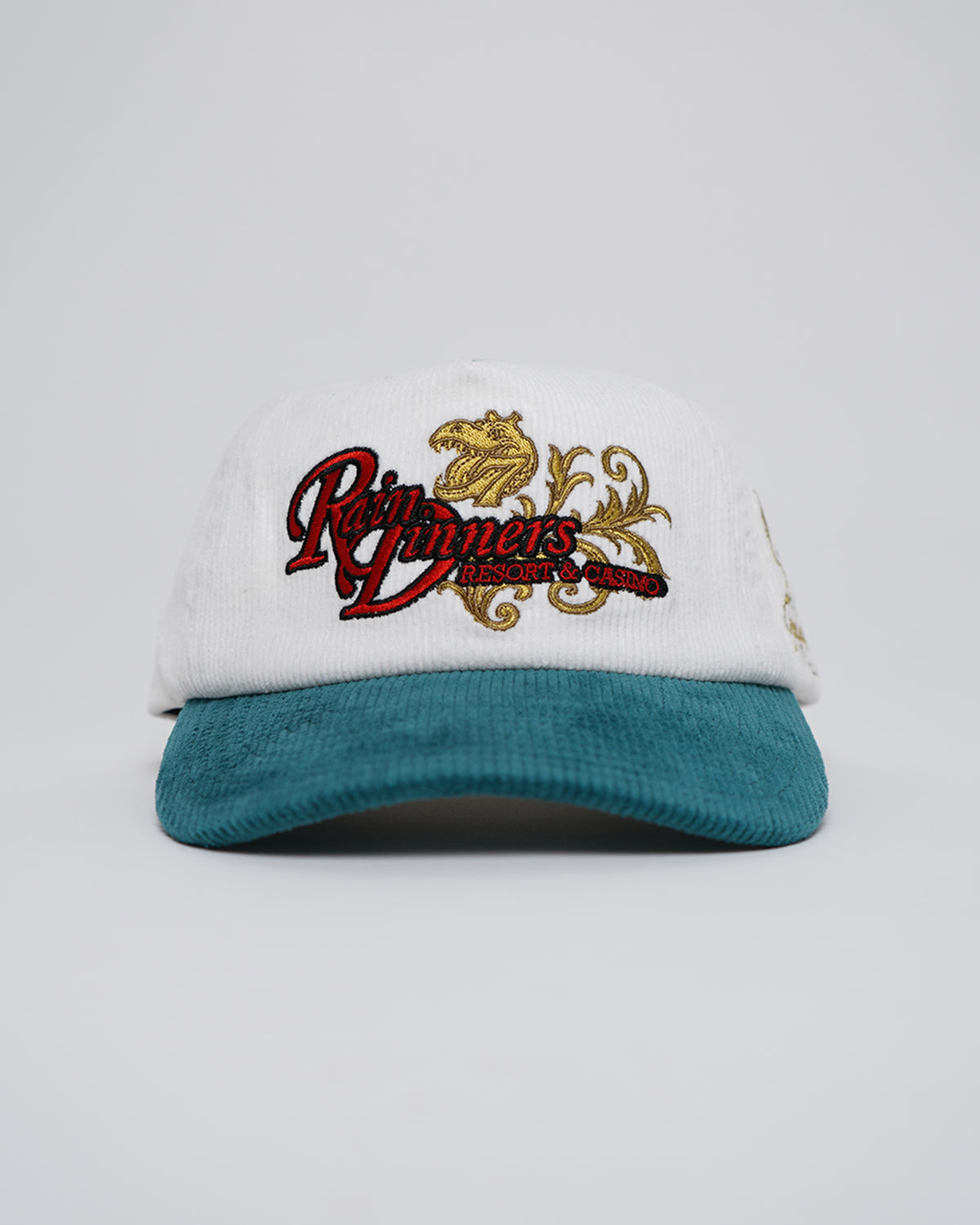 Front view of the Rain Dinners Hat, a white corduroy cap with teal brim, red script embroidery, and a gold dinosaur head with scrollwork