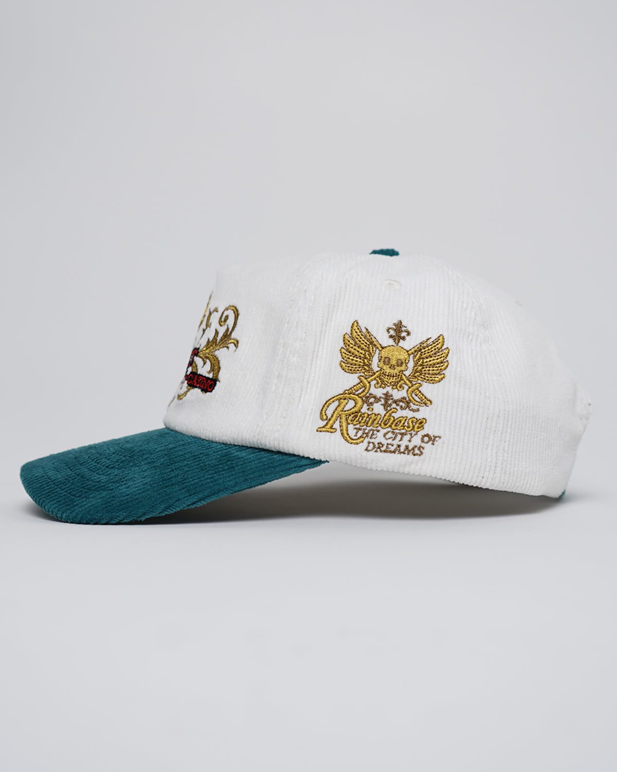 Side view of the Rain Dinners Hat, a white and teal corduroy cap with gold embroidered winged skull graphic and text