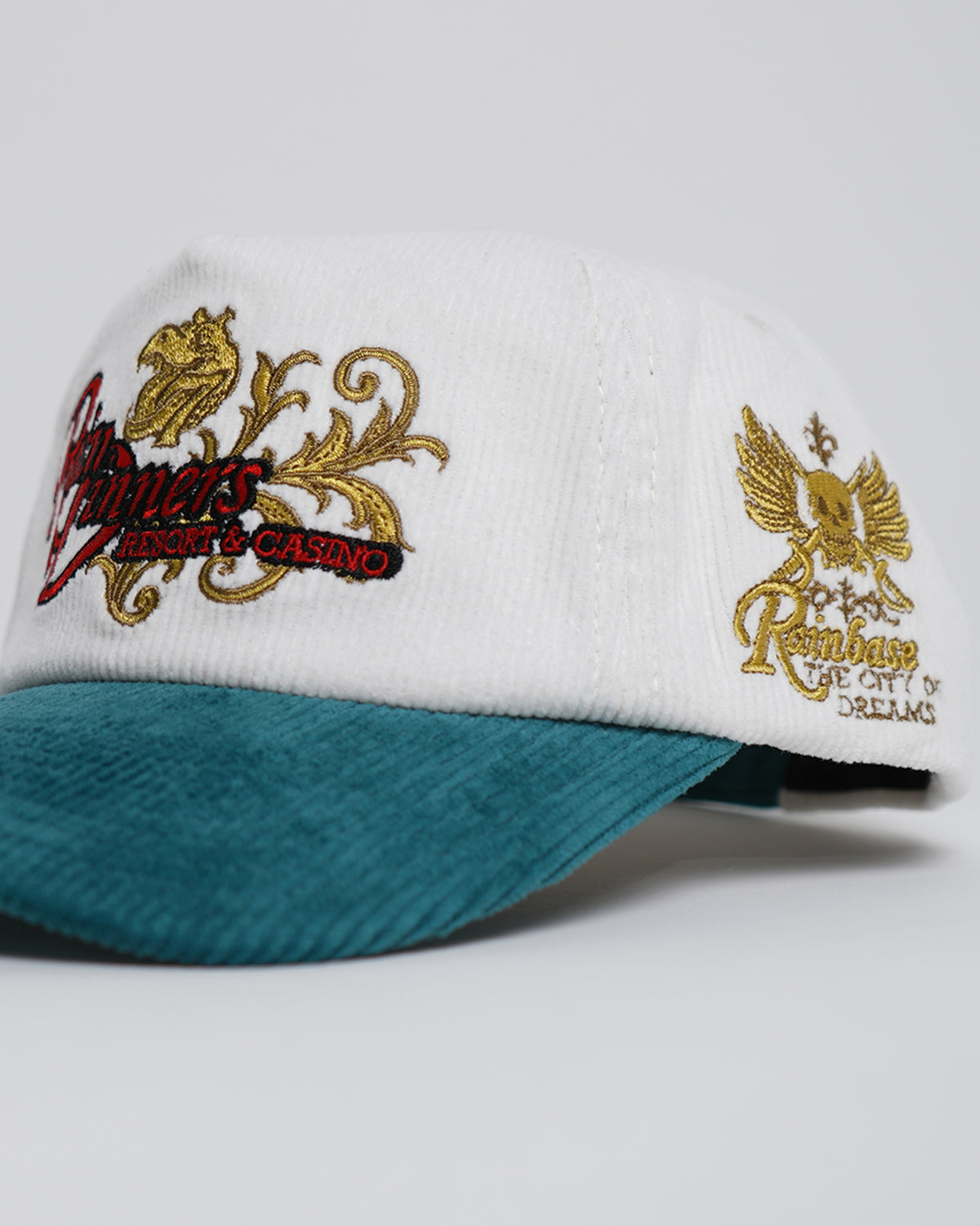 Side close-up of the Rain Dinners Hat, a white corduroy cap with teal brim, gold and red embroidered script logos and a winged skull emblem