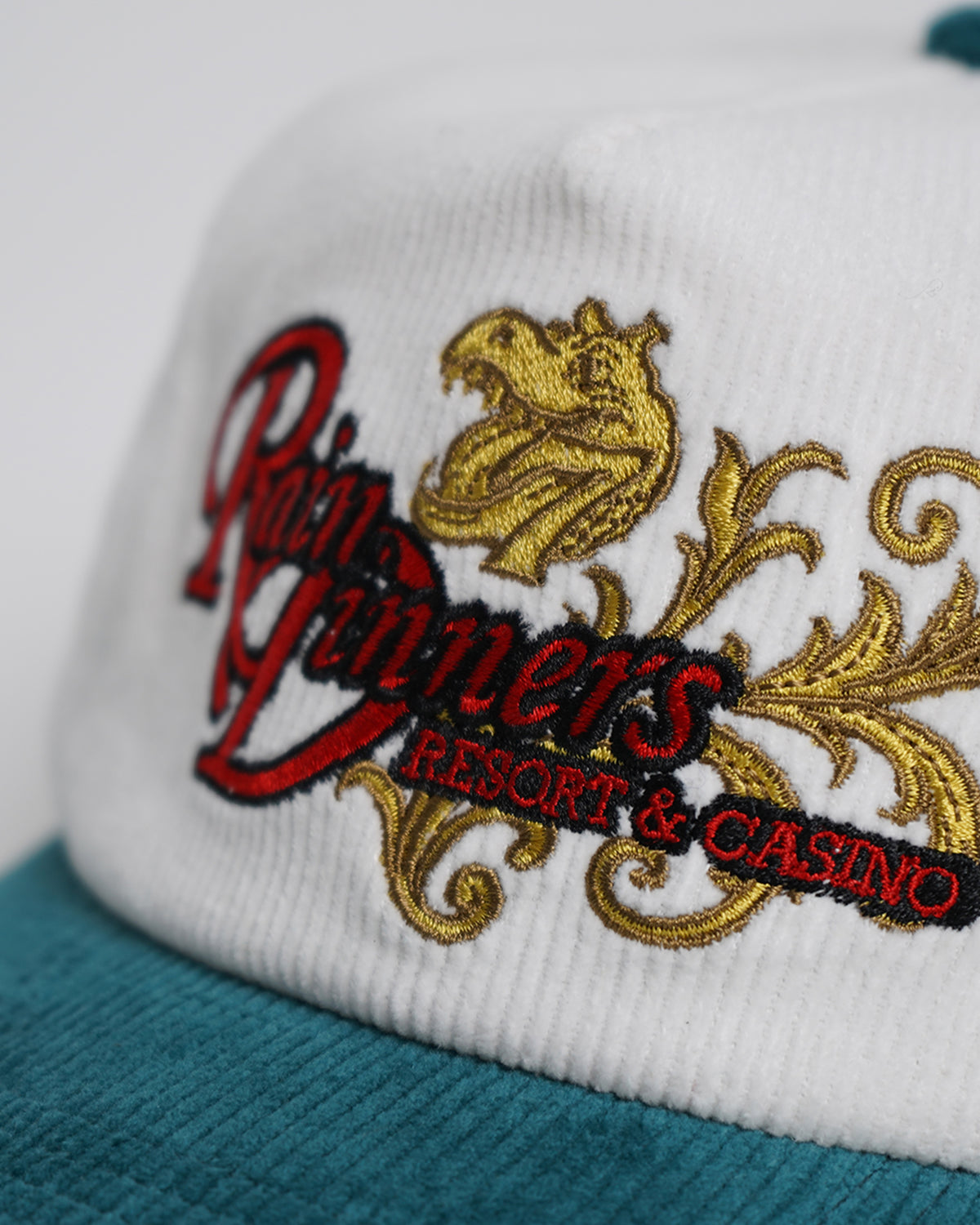 Close-up of the Rain Dinners Hat's white corduroy front panel and teal brim, with red embroidered script text, gold ornate scrollwork and a gold creature head