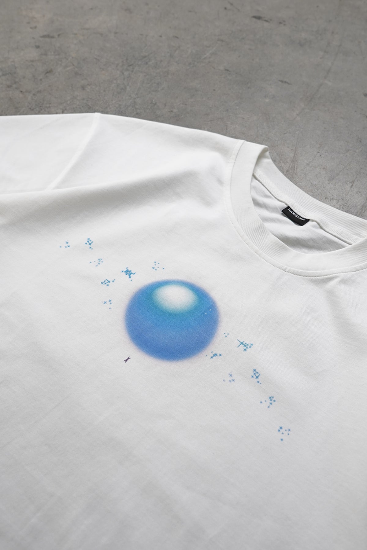 Close-up flat lay of the white Spirit Bomb Tee showing a blurry blue orb graphic with small blue sparkles around it and the black neck label