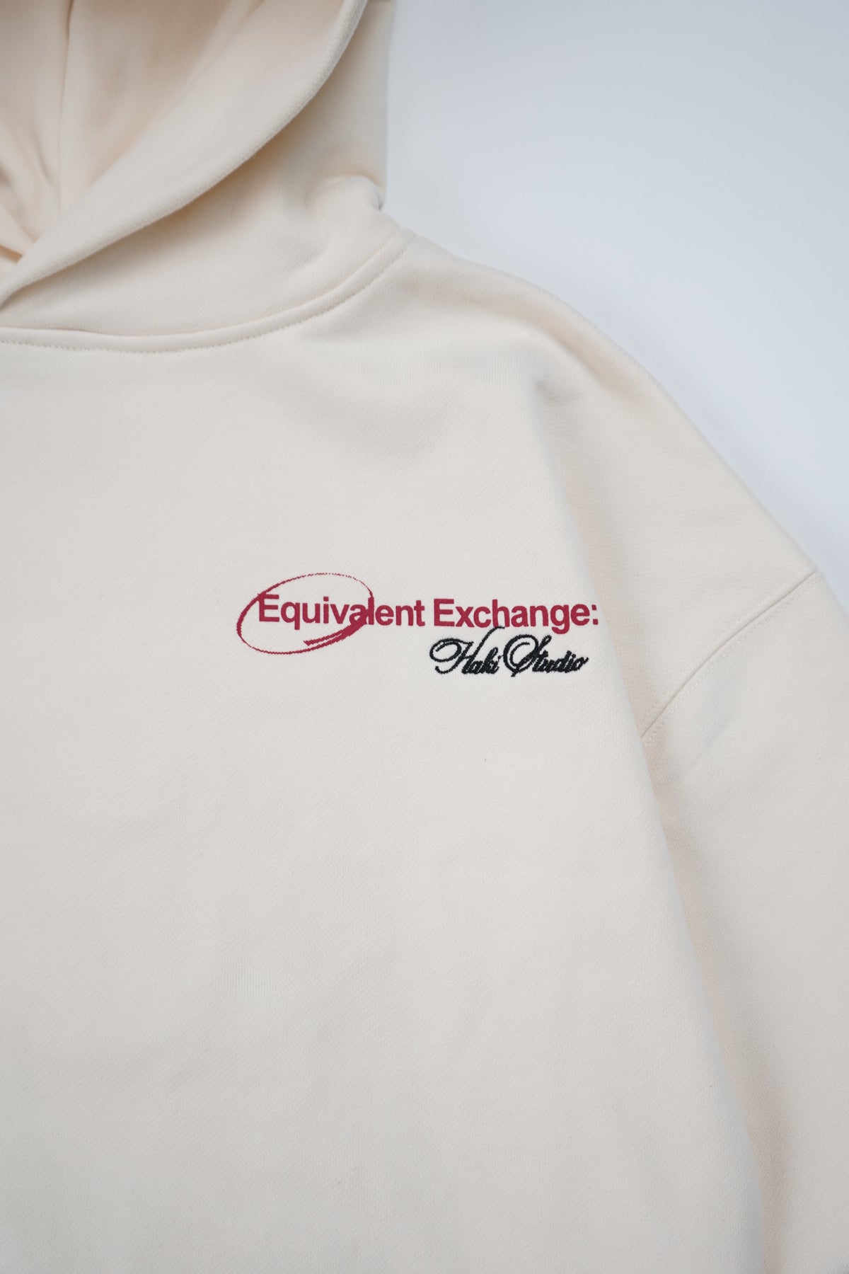 A close-up of a cream-colored hoodie featuring a red and black embroidered logo on the chest that reads 'Equivalent Exchange'.