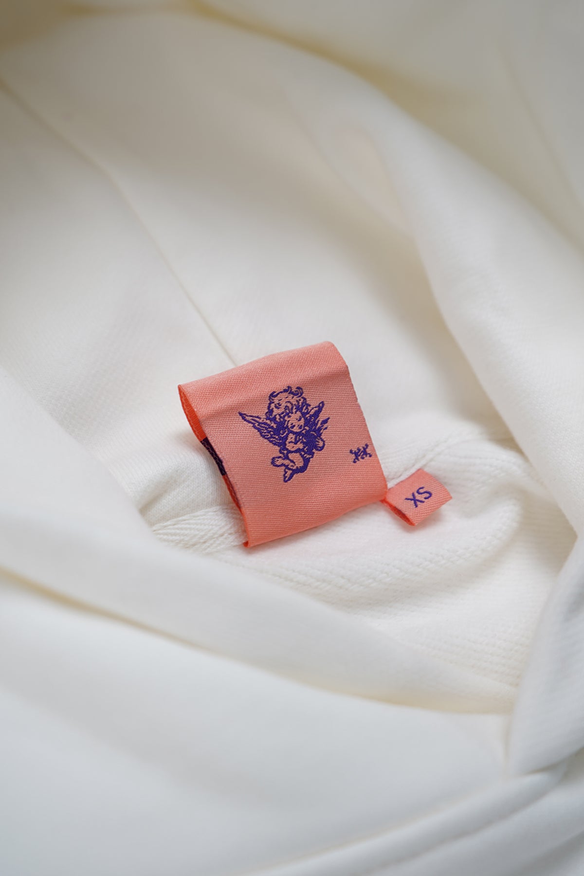 Close-up of a coral pink woven label with a purple cherub graphic and an XS size tab, inside the cream white Together Forever Hoodie