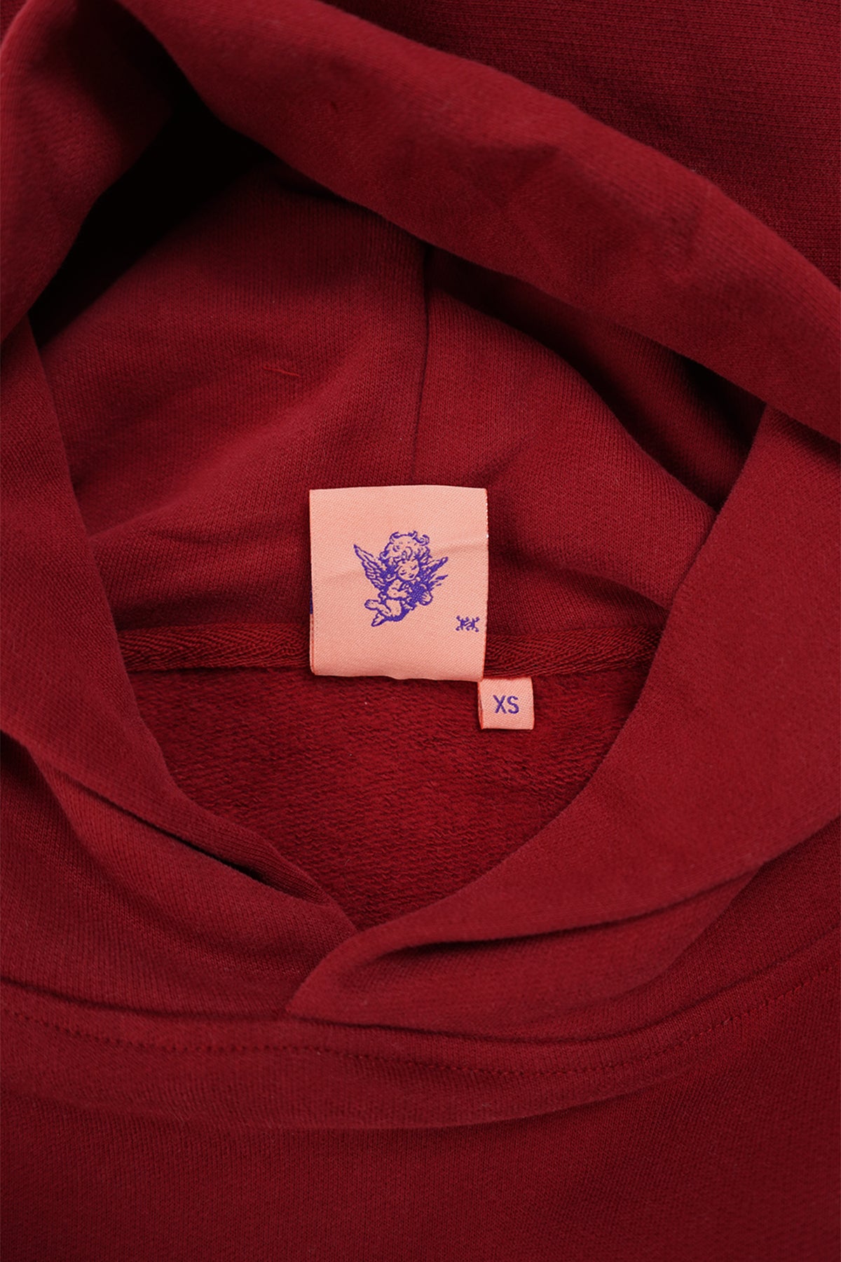 Close-up of the Cherry Red Come Over Hoodie interior at the neck, showing a pink woven label with a purple cherub graphic and an XS size tag