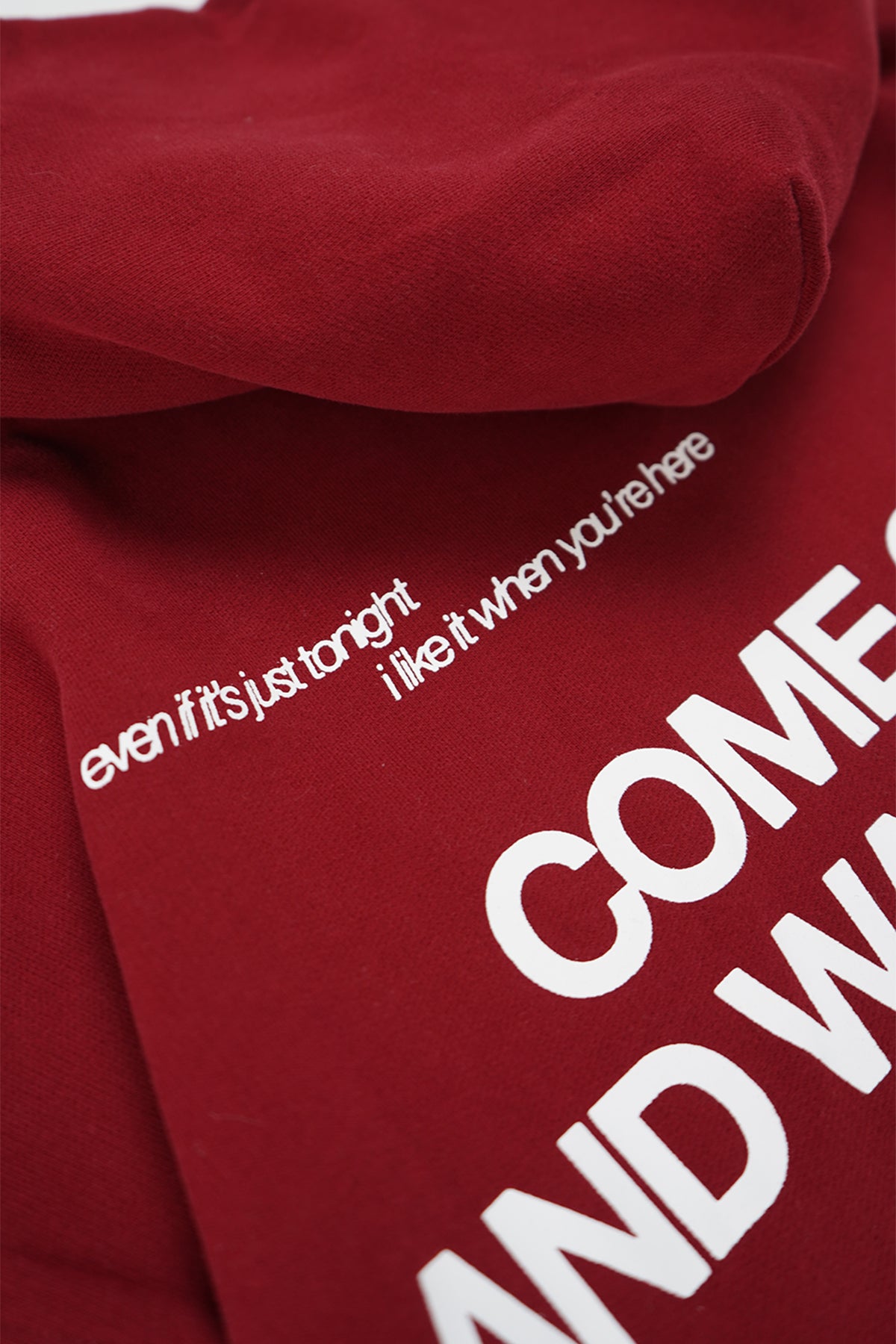 Close-up of the Cherry Red Come Over Hoodie showing small white printed text reading "even if it's just tonight i like it when you're here" above large partly visible white block letters