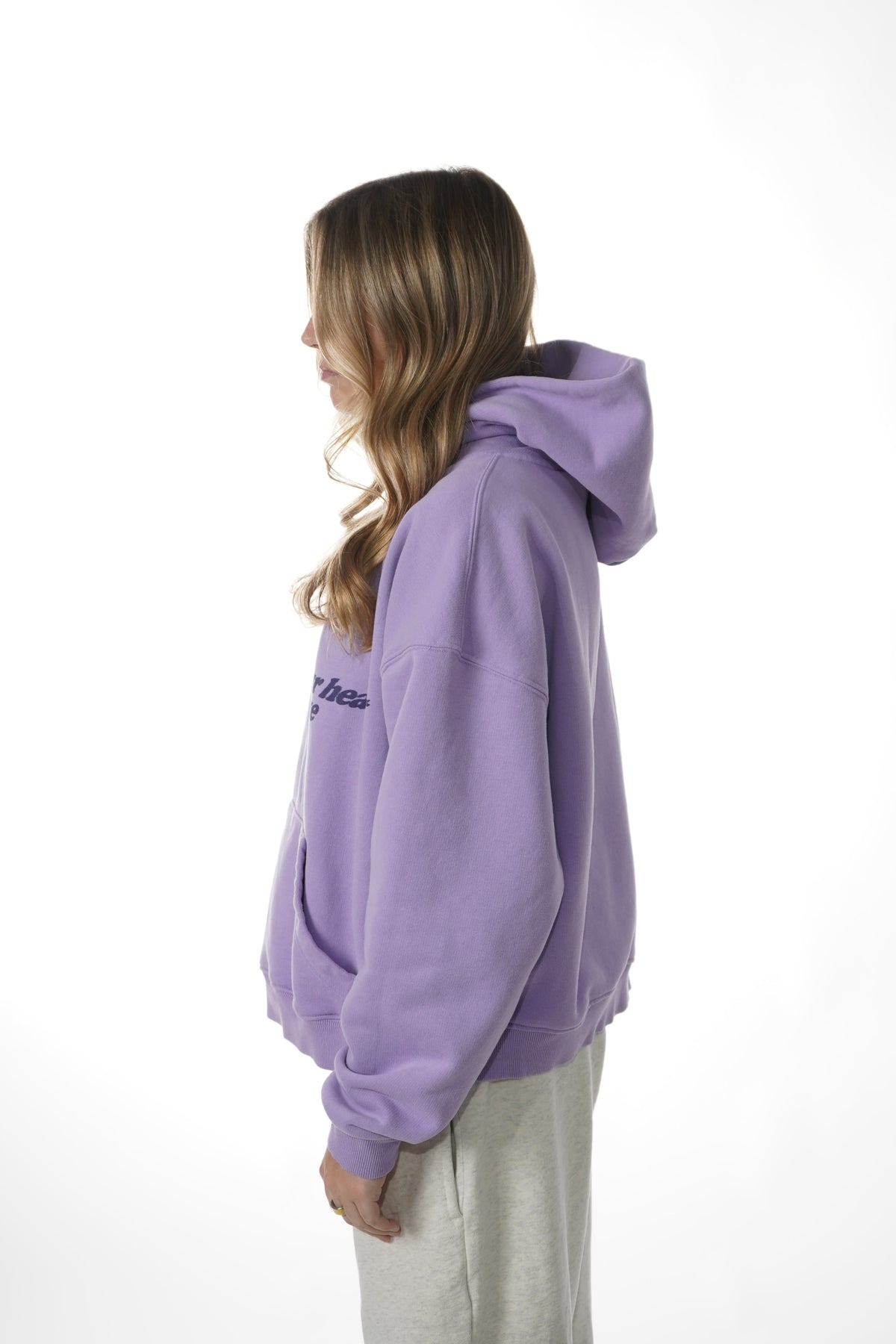 A woman shown from the side profile wearing a lavender purple pullover hoodie with a small embroidered logo on the chest, paired with light gray sweatpants, against a plain white studio background.
