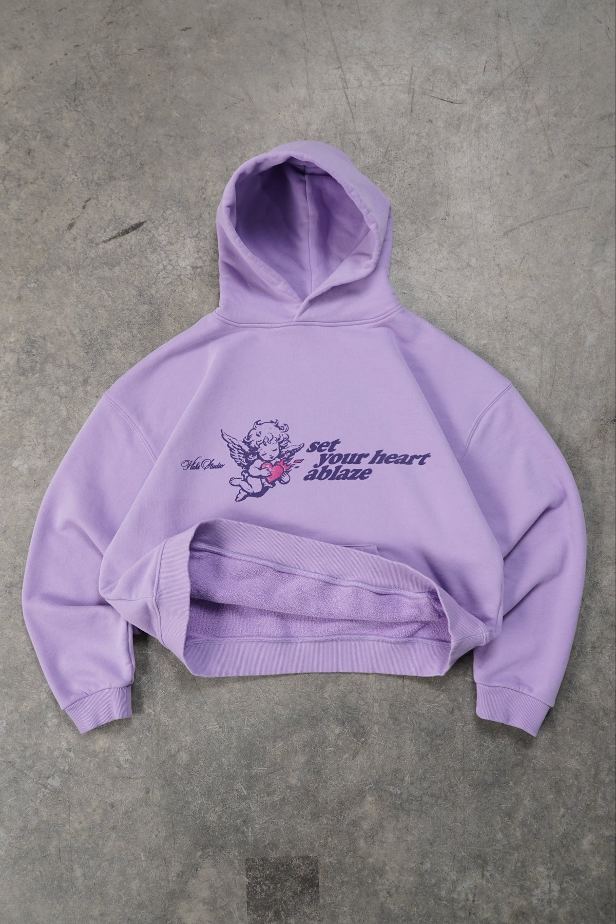 A lavender purple cropped hoodie with a graphic print on the front, laid flat on a textured gray concrete floor. The graphic features a small cartoon character holding a pink heart and the text 'get your heart above ground'.