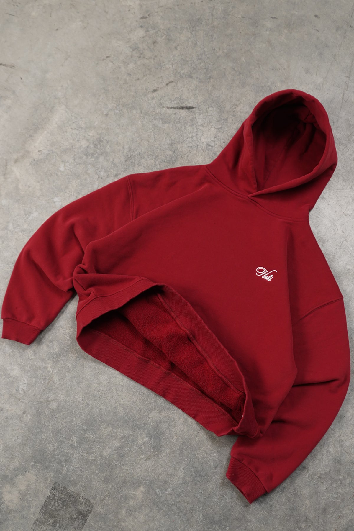 Flat lay of the Cherry Red Come Over Hoodie on concrete, front view with small white script embroidery on the chest and the red fleece interior visible at the hem