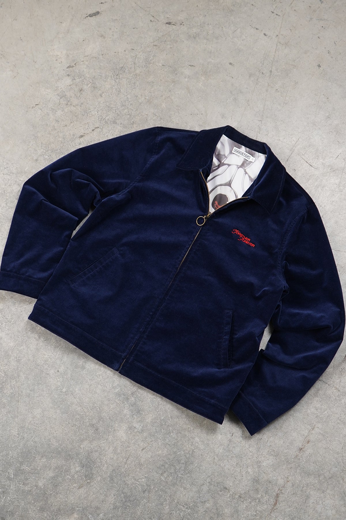 Flat lay of the navy corduroy Queen of Curses Jacket, front view, with collar, front zip, side pockets, red embroidered script on the chest and a grey printed lining