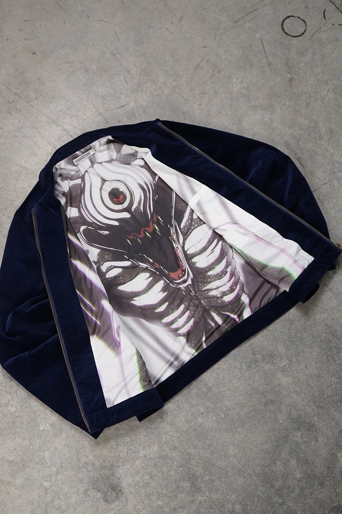 Flat lay of the open navy corduroy Queen of Curses Jacket with gold zipper, showing the interior lining printed with a one-eyed skeletal creature graphic