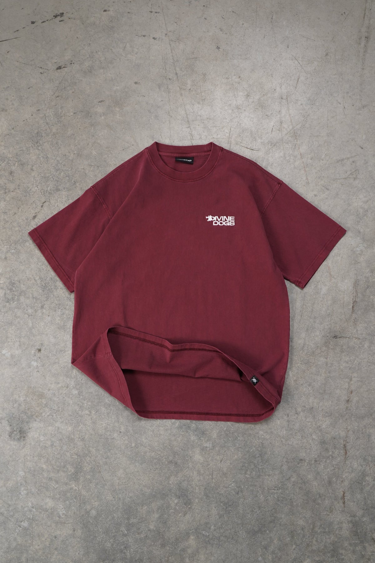 A maroon short-sleeve t-shirt with a small white graphic on the chest, laid flat on a textured gray concrete floor.