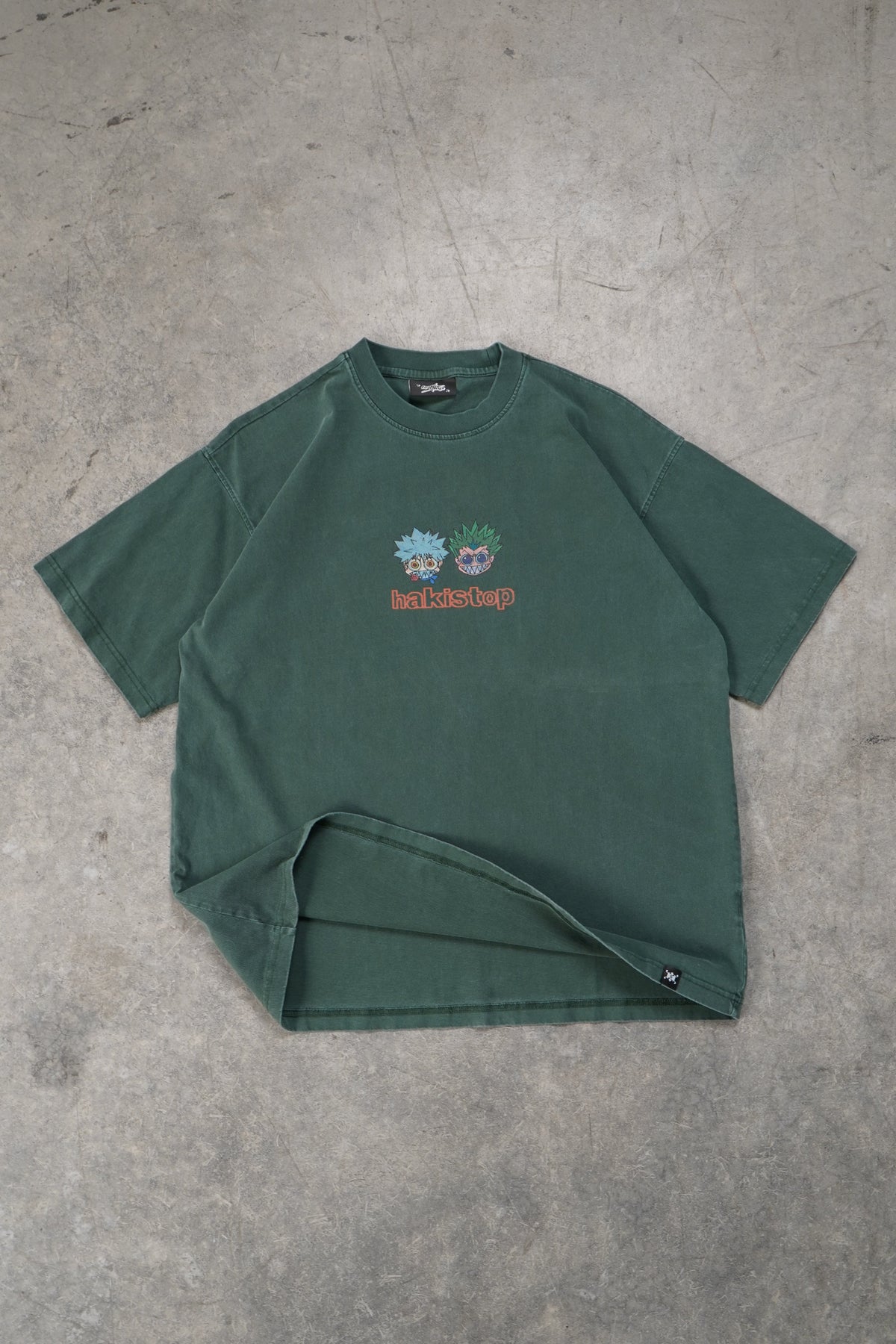 A dark green short-sleeve t-shirt with a small colorful chest graphic, laid flat on a textured gray concrete floor. The graphic features a blue and green character with the word 'hakistop' printed below it.