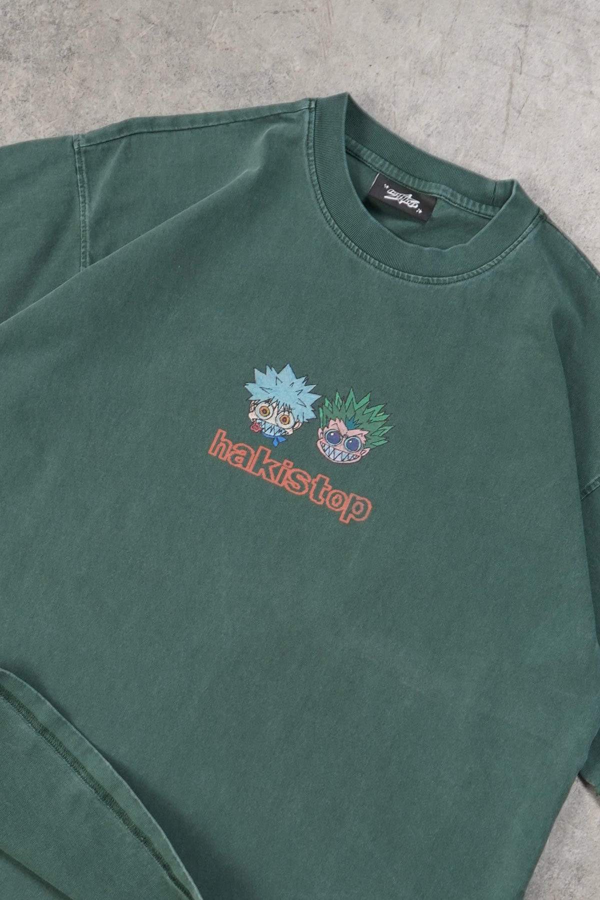 A dark green short-sleeve t-shirt with a small colorful chest graphic, laid flat on a textured gray concrete floor.