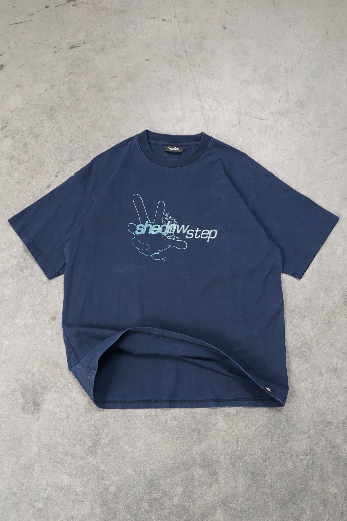 A navy blue short-sleeve t-shirt with a white graphic print on the front, laid flat on a textured gray concrete floor. The graphic features a rabbit silhouette and the text 'the shadow step'.