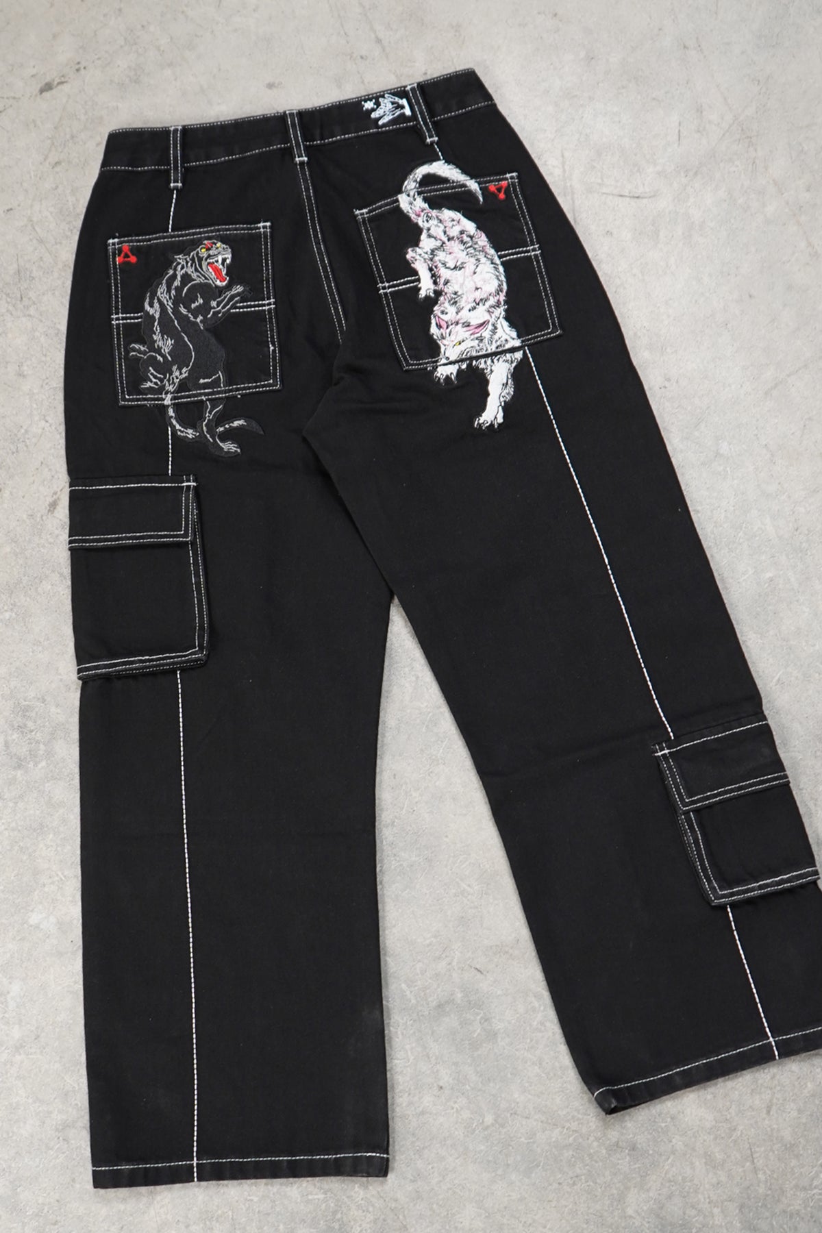 Flat lay back view of the black Divine Dogs Pants with white contrast stitching, embroidered black panther and white wolf on back pockets, and side cargo pockets