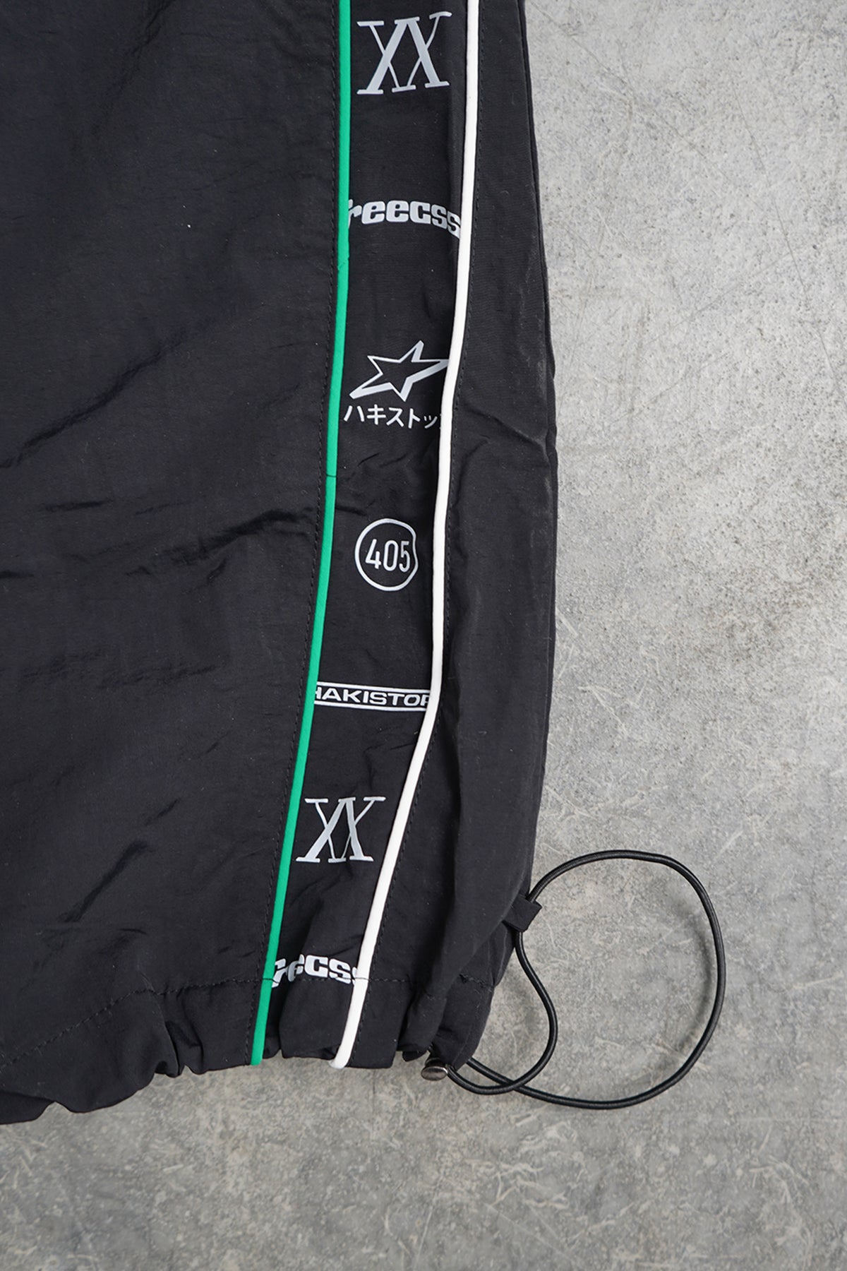 Close-up of the Jan-ken Trackpants black nylon hem with green and white piping, printed side tape of logos and numerals, and a black elastic drawcord