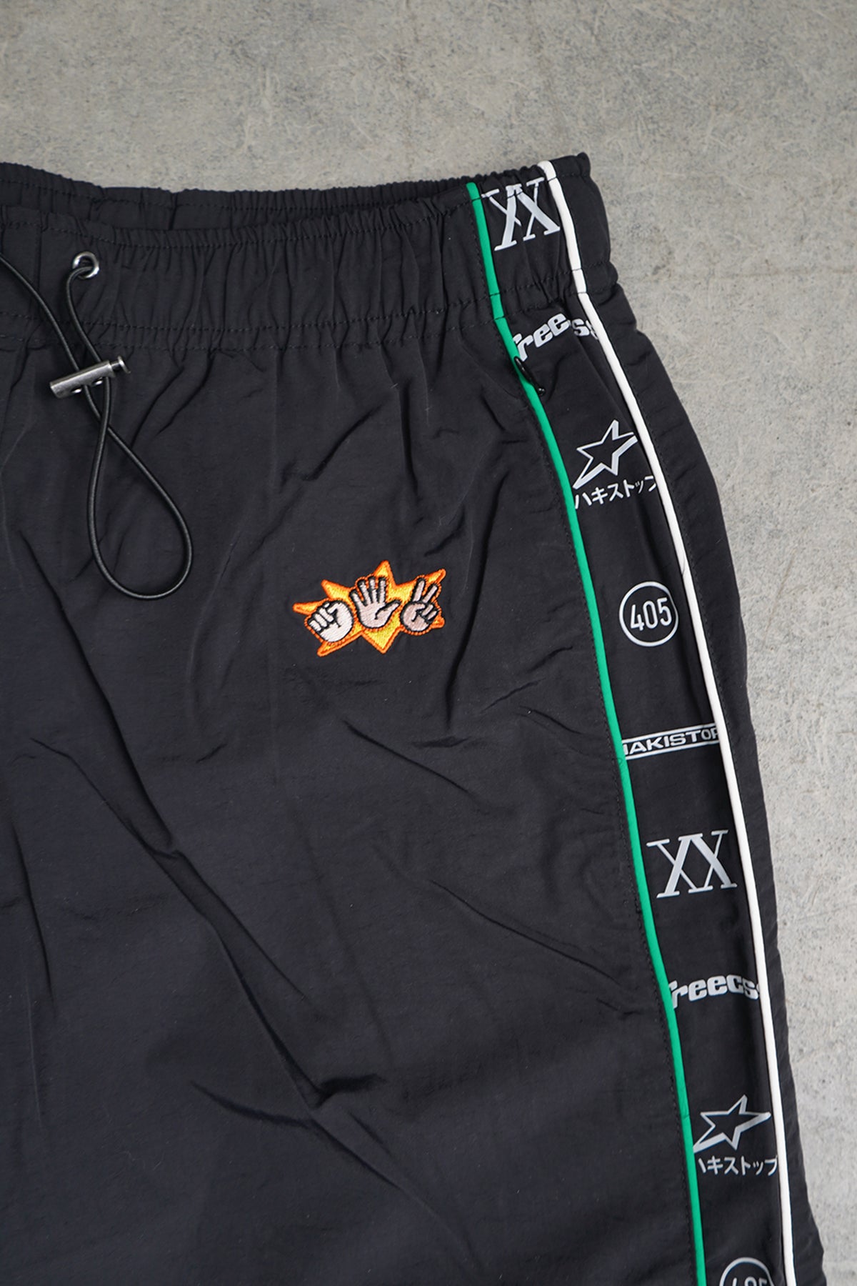 Close-up of the black Jan-ken Trackpants waistband and hip, with drawstring, embroidered rock-paper-scissors hands on an orange burst, and side tape with green and white piping