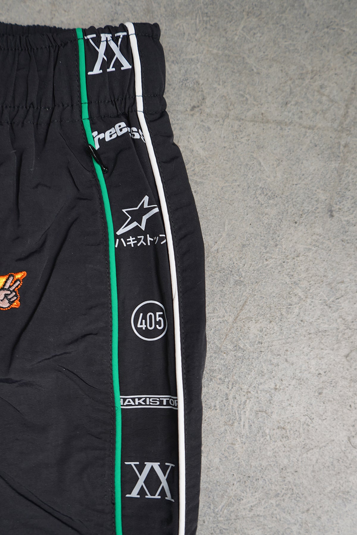 Close-up of the Jan-ken Trackpants in black, showing the elastic waistband, green and white side piping, and a side stripe of printed Roman numeral XX, a star and Japanese text